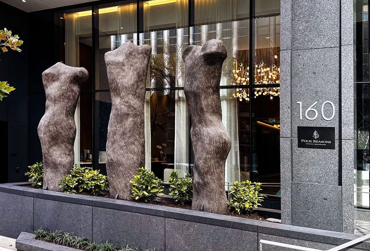 Three large, sculptural wooden logs displayed vertically in a modern planter outside a building with large glass windows. The building has the number 160 and a sign that reads Four Seasons.