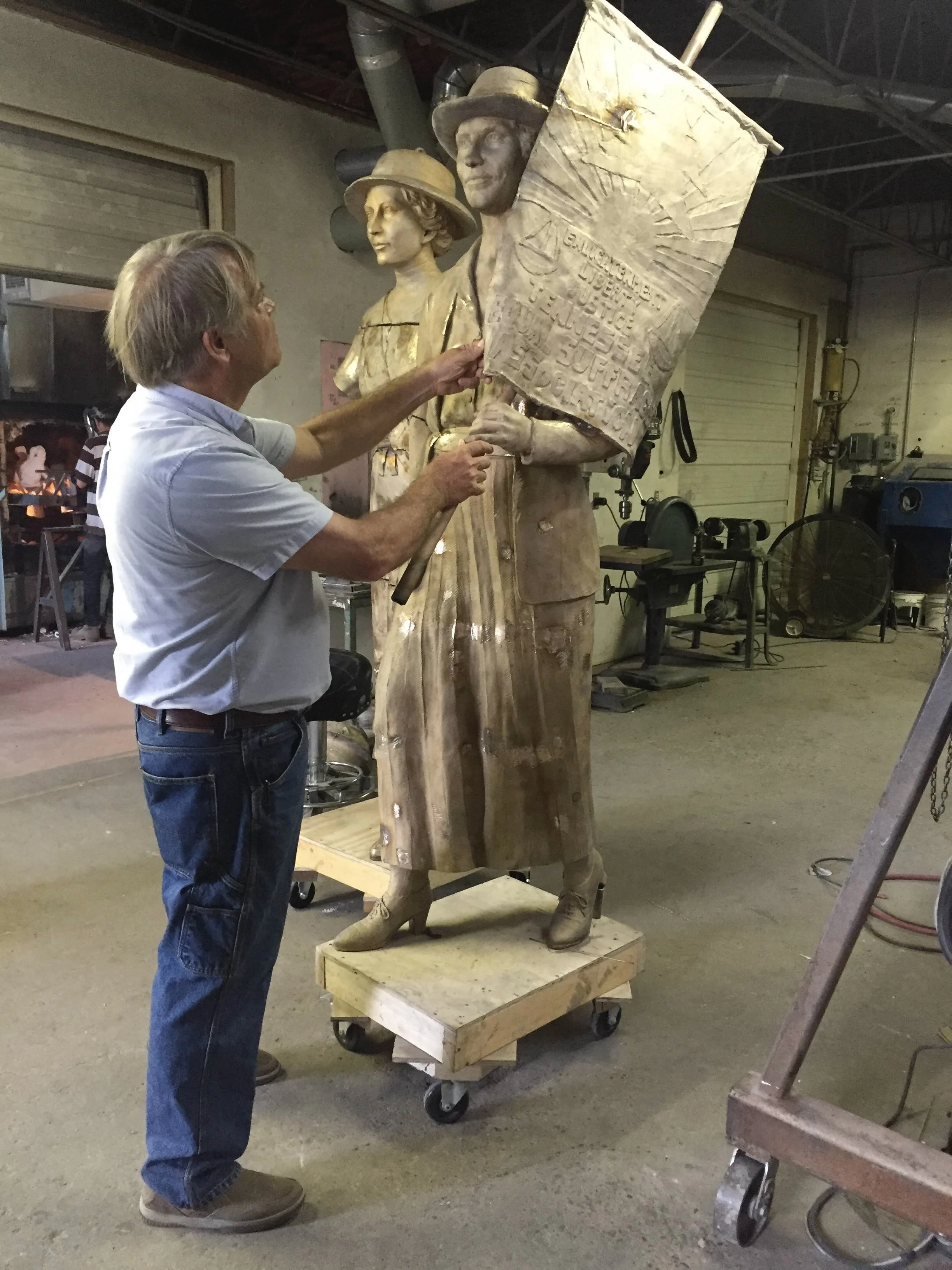 A man working on a large bronze sculpture of two people, woman, both wearing hats. The sculpture is on a wheeled platform and the woman is holding a sign with inscriptions. Woman's Suffrage. 