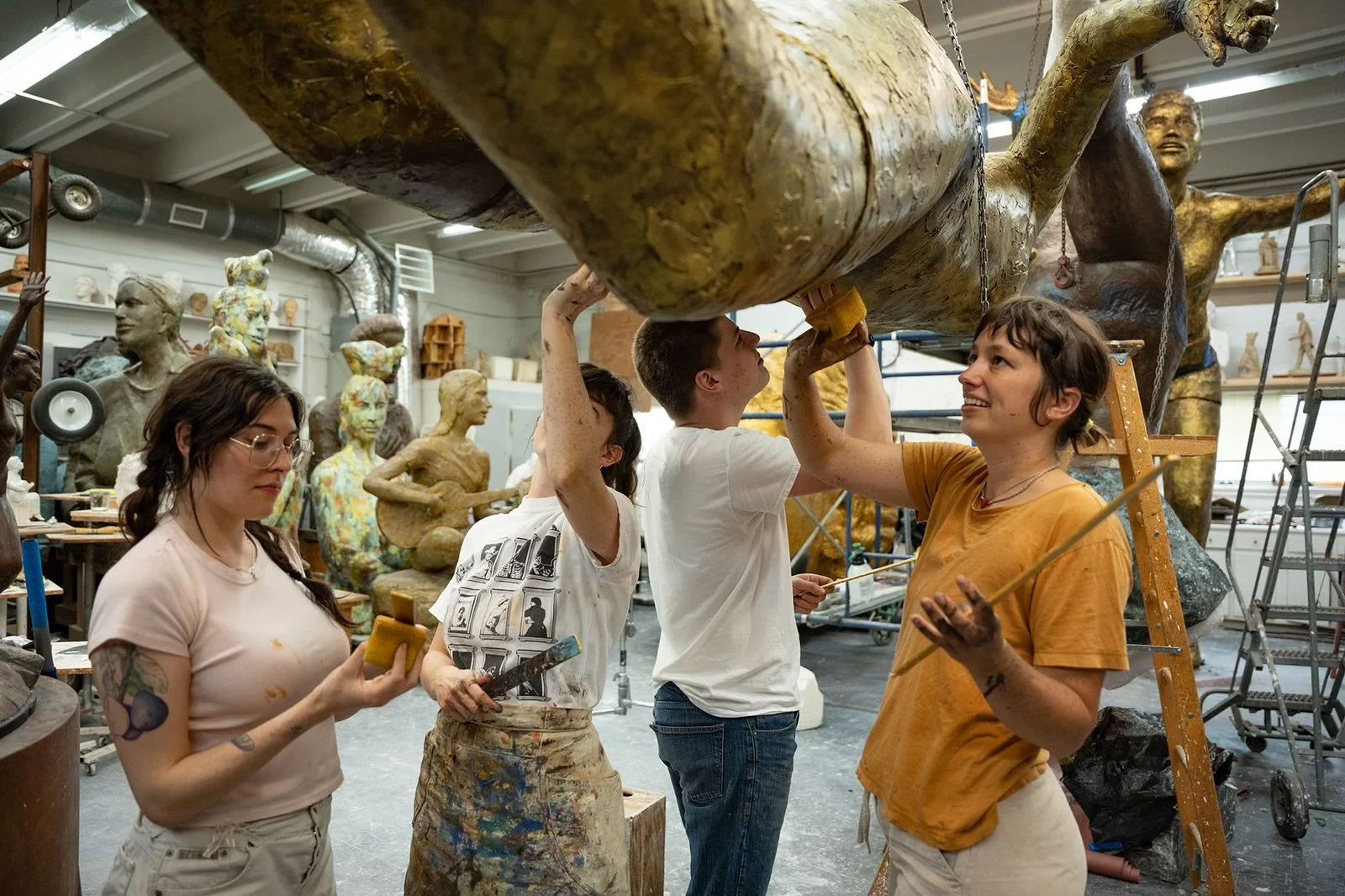 A group of artists working together in a sculpture studio, with one person holding a sponge, another painting, and two lifting a large sculpture.