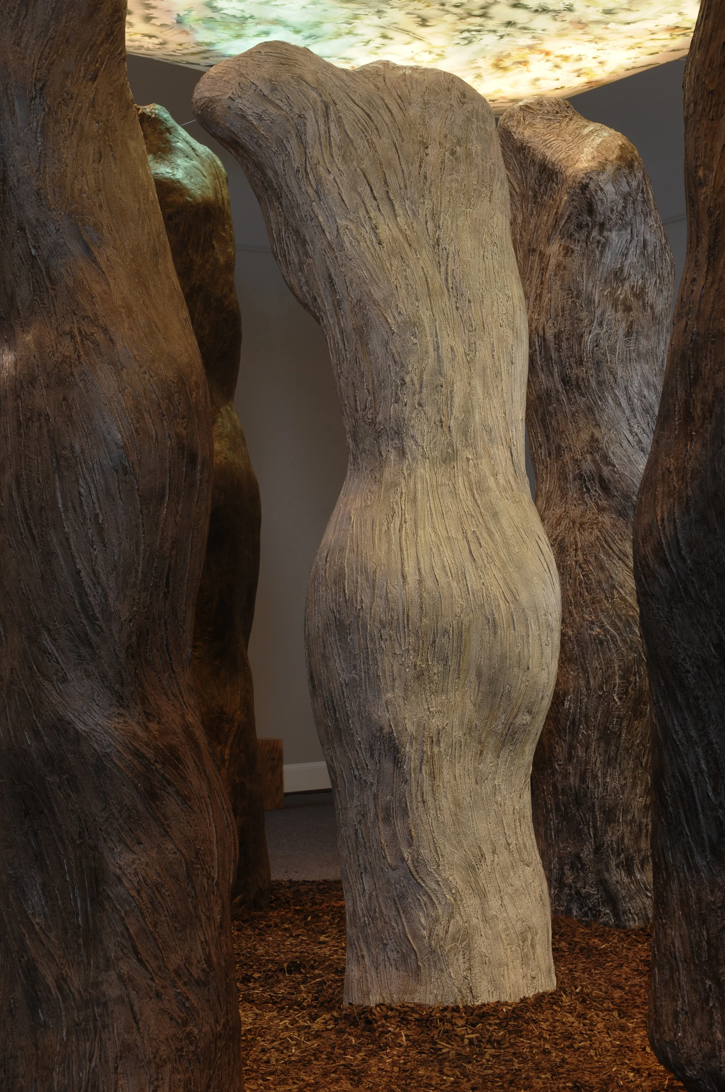 Close-up of large, textured wooden sculptures resembling tree trunks with a painted, cloud-like ceiling overhead and wooden chips on the floor.