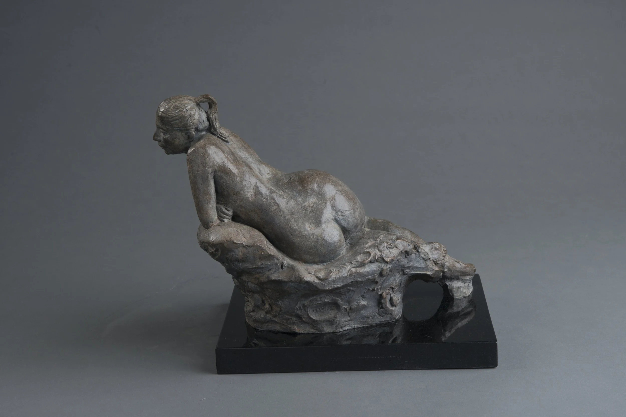 A bronze sculpture of a woman lying on her side, with her head resting on her arm, on a rocky base, against a plain grey background.