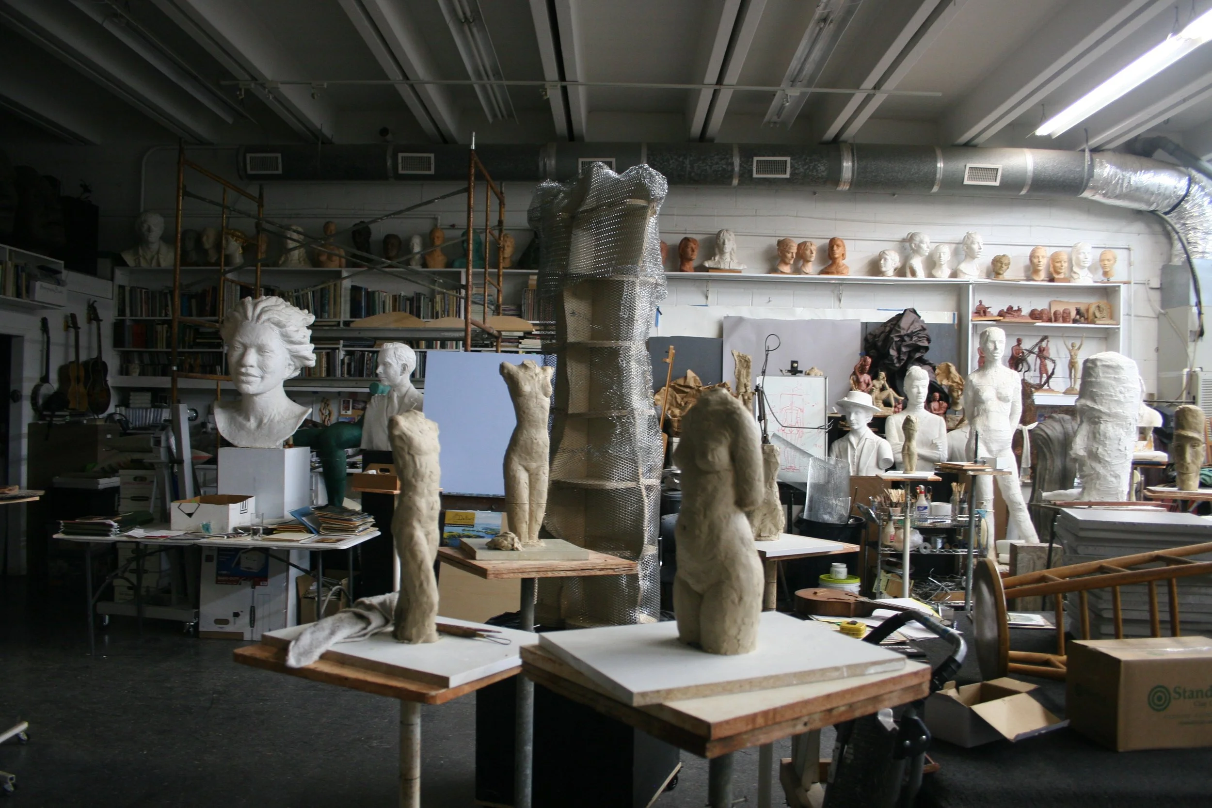 An artist's studio filled with sculpture busts, sketches, books, and art supplies, with large ductwork and fluorescent lights overhead.