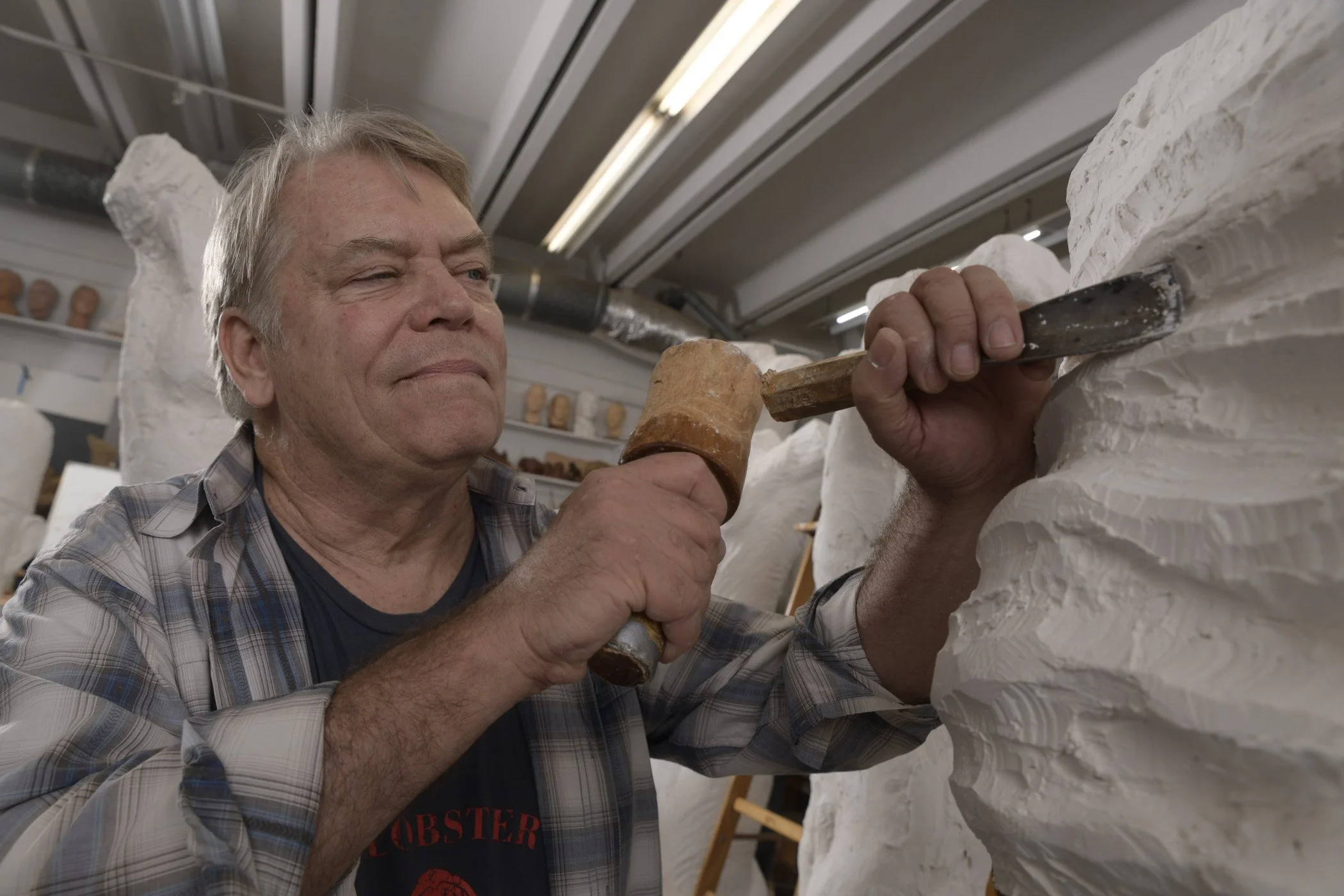 A man with gray hair wearing a plaid shirt and a lobster-themed shirt, working on a white sculpted wall using a hammer and chisel in an art studio.