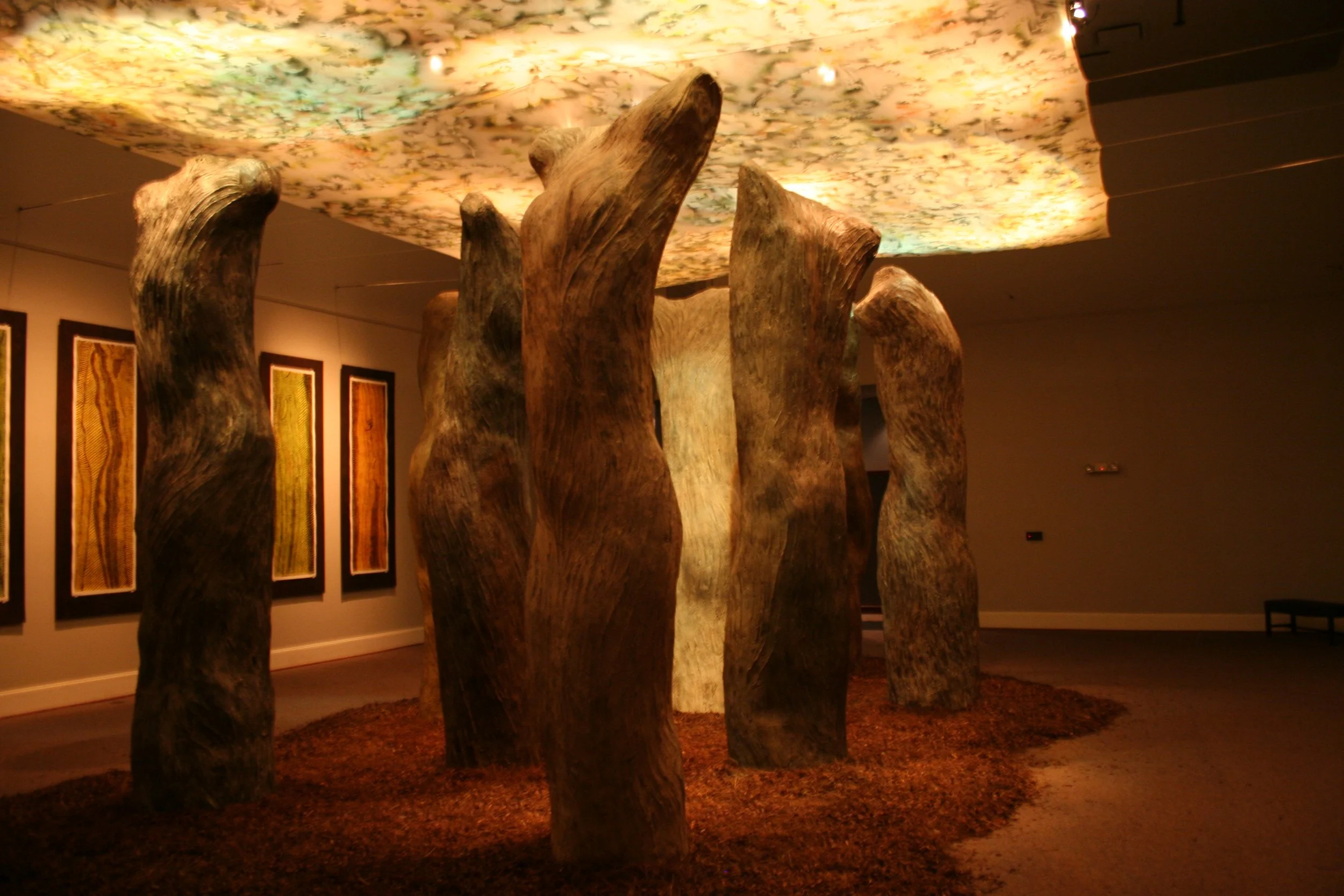 Art installation featuring large wooden sculptural forms resembling tree trunks, displayed in an indoor gallery with framed artwork on the walls and a ceiling decorated with a floral pattern. Dream Forest Installation.