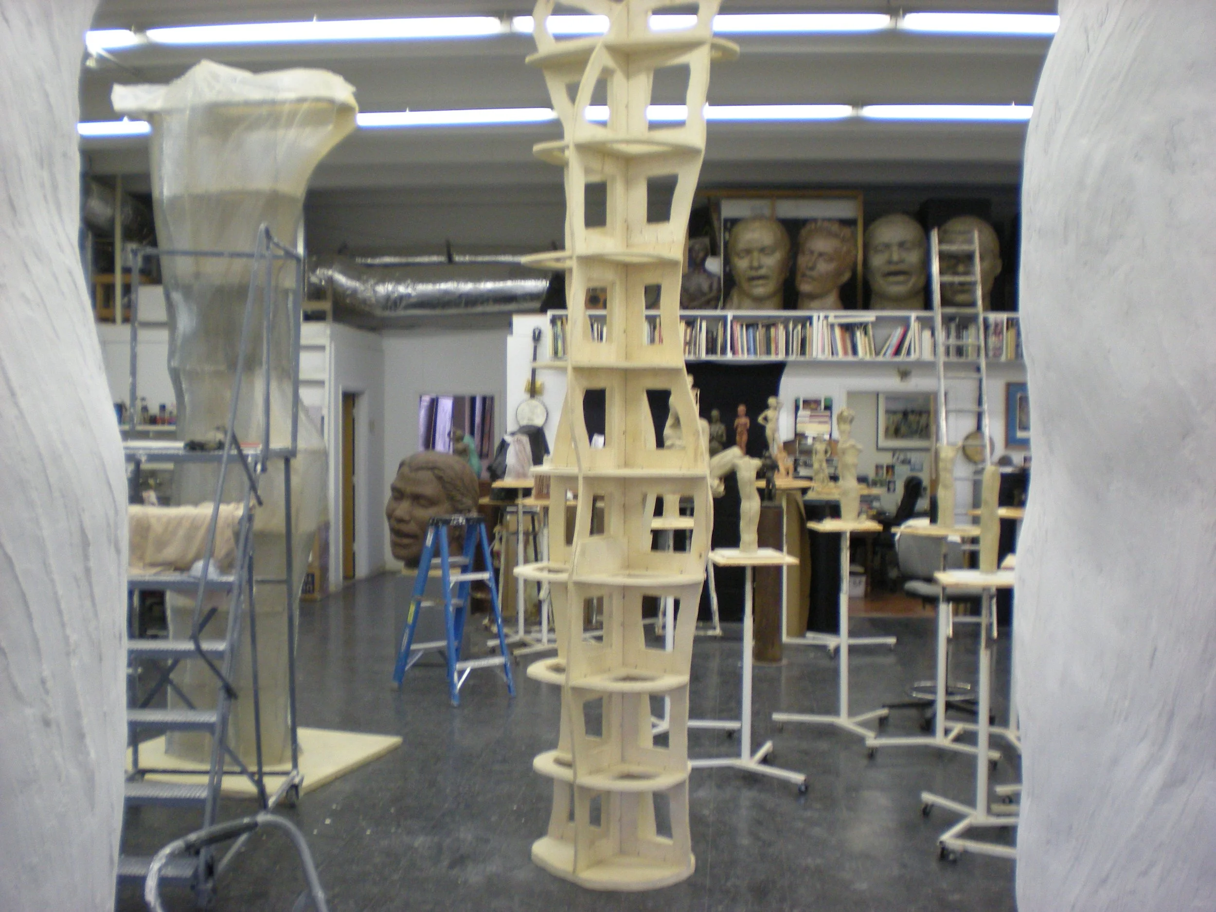 View through two white textured sculptures into an art studio or gallery space with shelves, bust sculptures, and a tall wooden sculpture structure in the center.