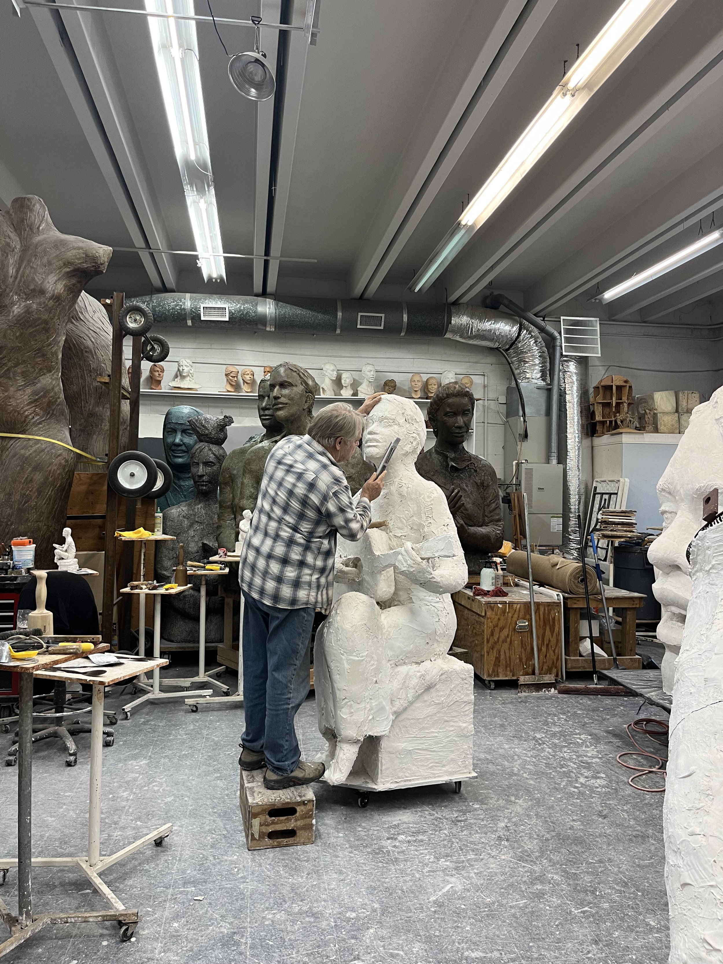 An artist in a workshop sculpting a large bust out of white material, with various other bust sculptures and art supplies around.
