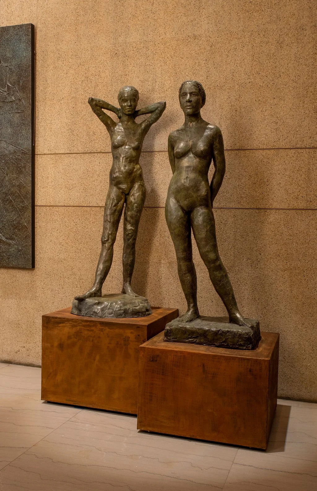 Two bronze sculptures of nude women on metal pedestals against a beige wall. One woman has her arms raised behind her head, and the other stands with hands behind her back.
