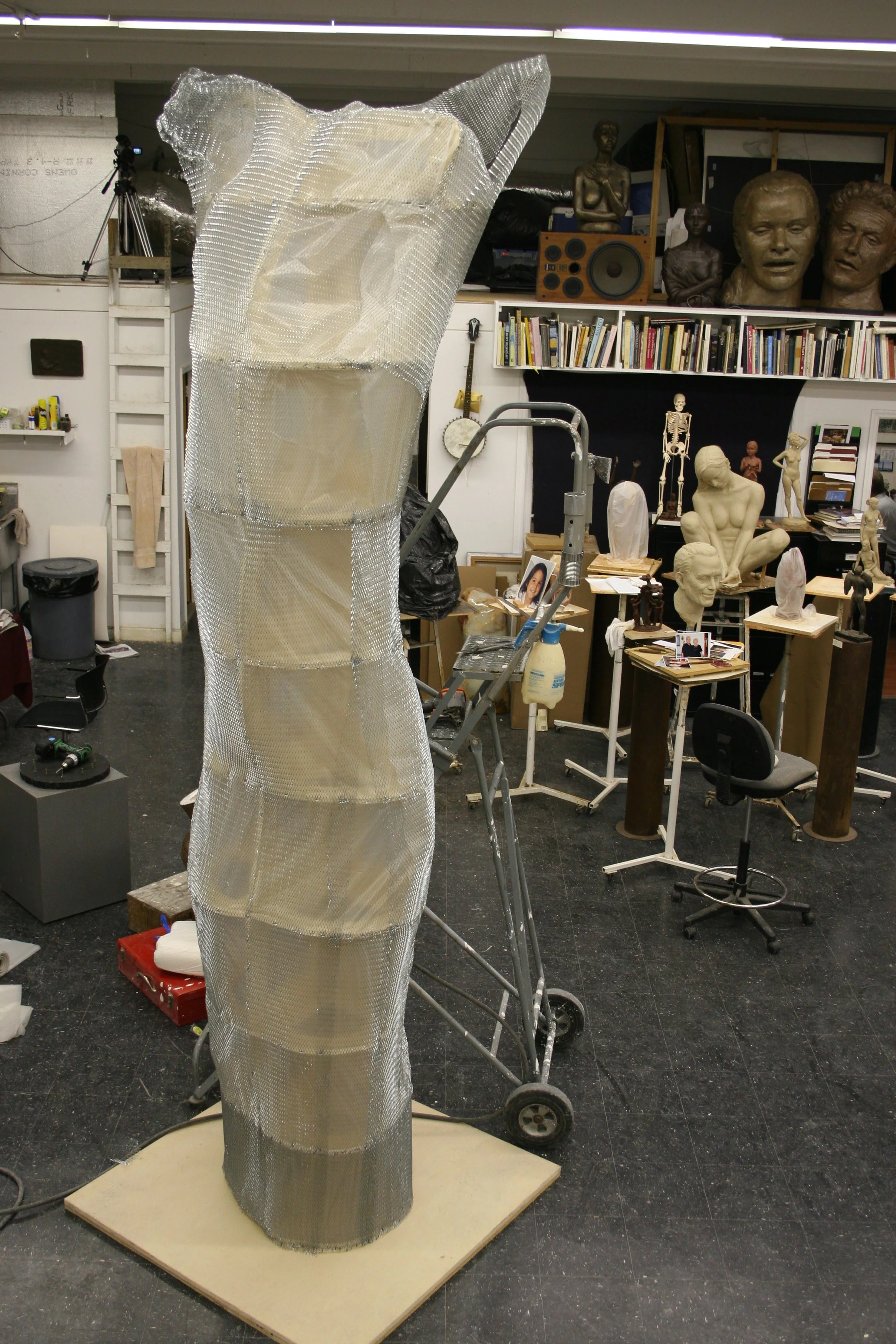 An art studio with various sculptures, books, and art supplies on shelves and tables. Prominent in the foreground is a tall sculpture covered in metal wrap. The background shows bust sculptures, a skeleton model, and audio equipment on shelves.