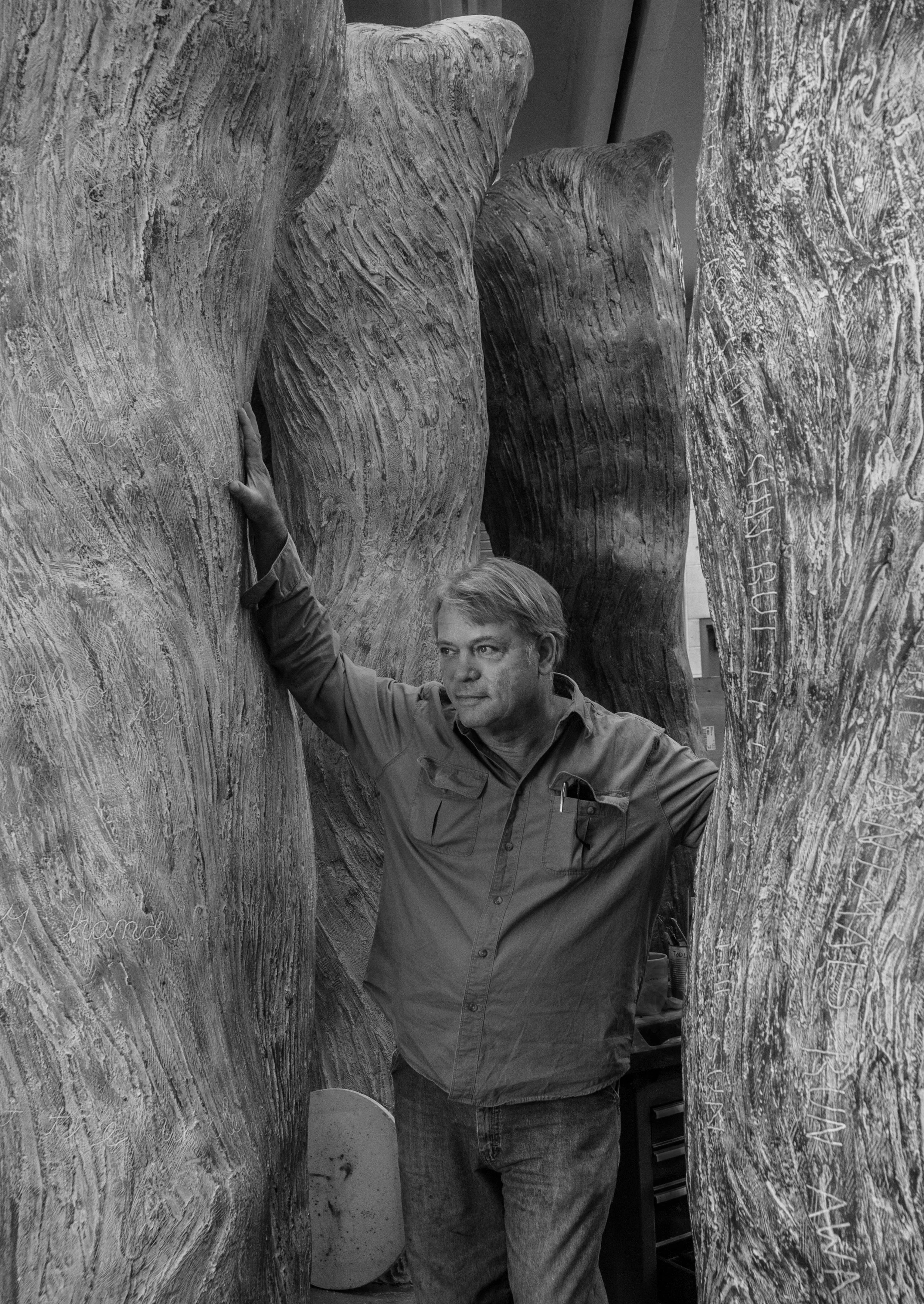 A man standing among large marble sculptures indoors, with one arm raised and touching a sculpture, in a workshop or gallery setting.