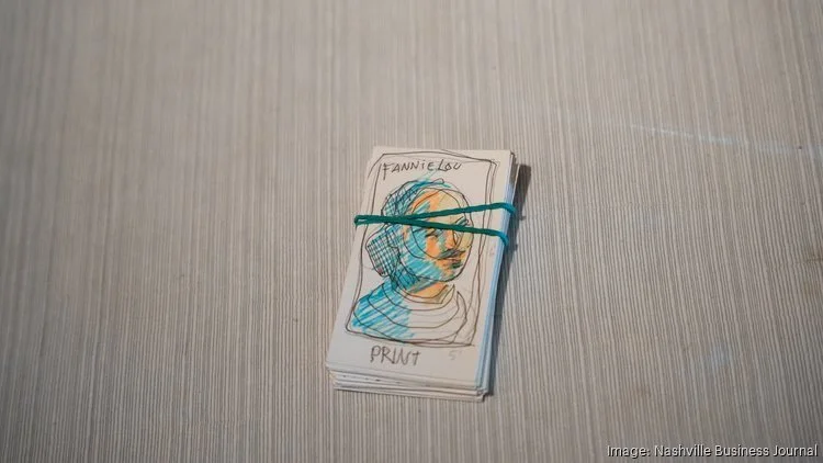 Stack of small paper notepads with a colorful sketch of a person's face on top, bound with a green rubber band, on a wooden surface.