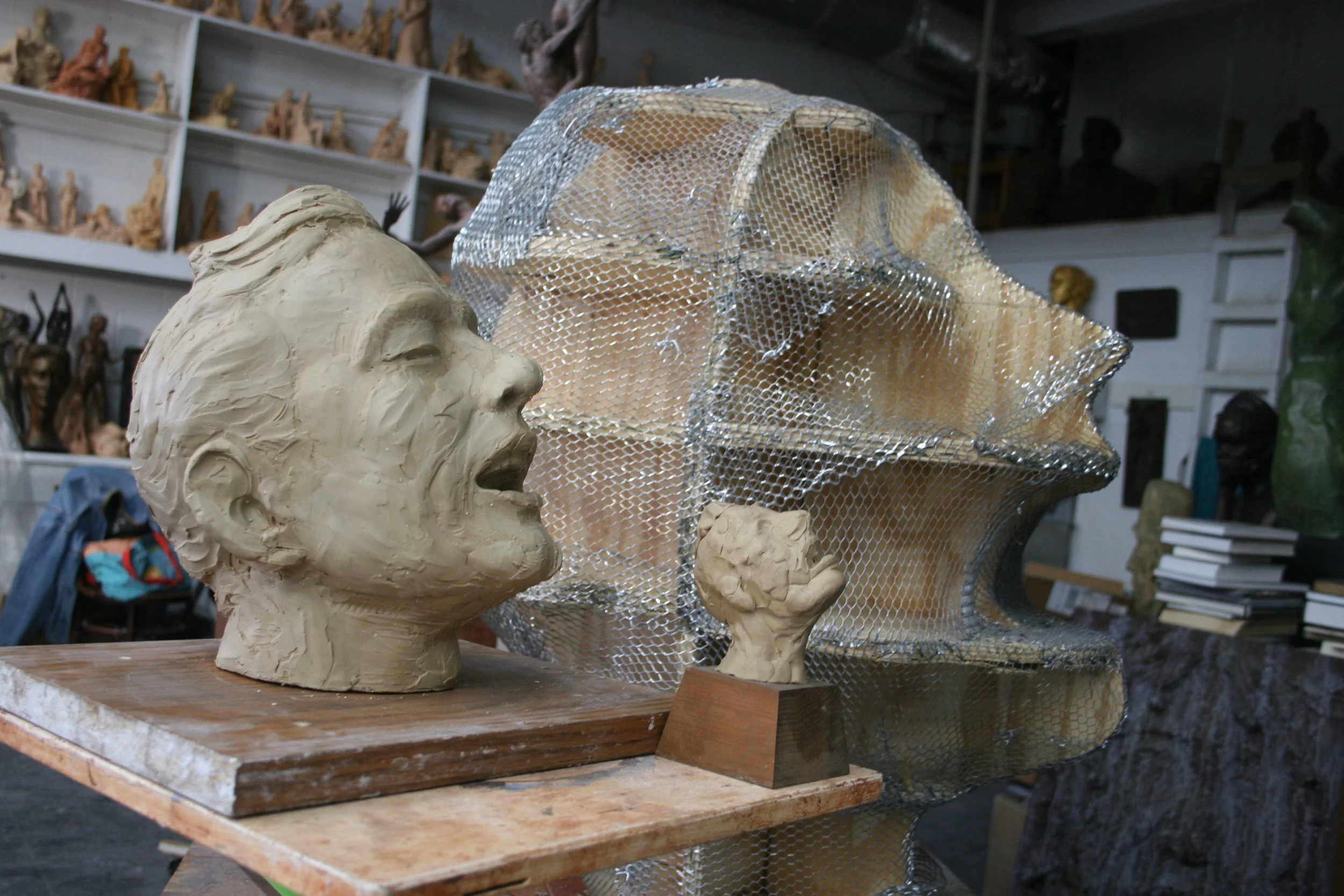 Clay busts of a young man and woman in an artist's studio, with shelves of small sculptures and art supplies in the background.