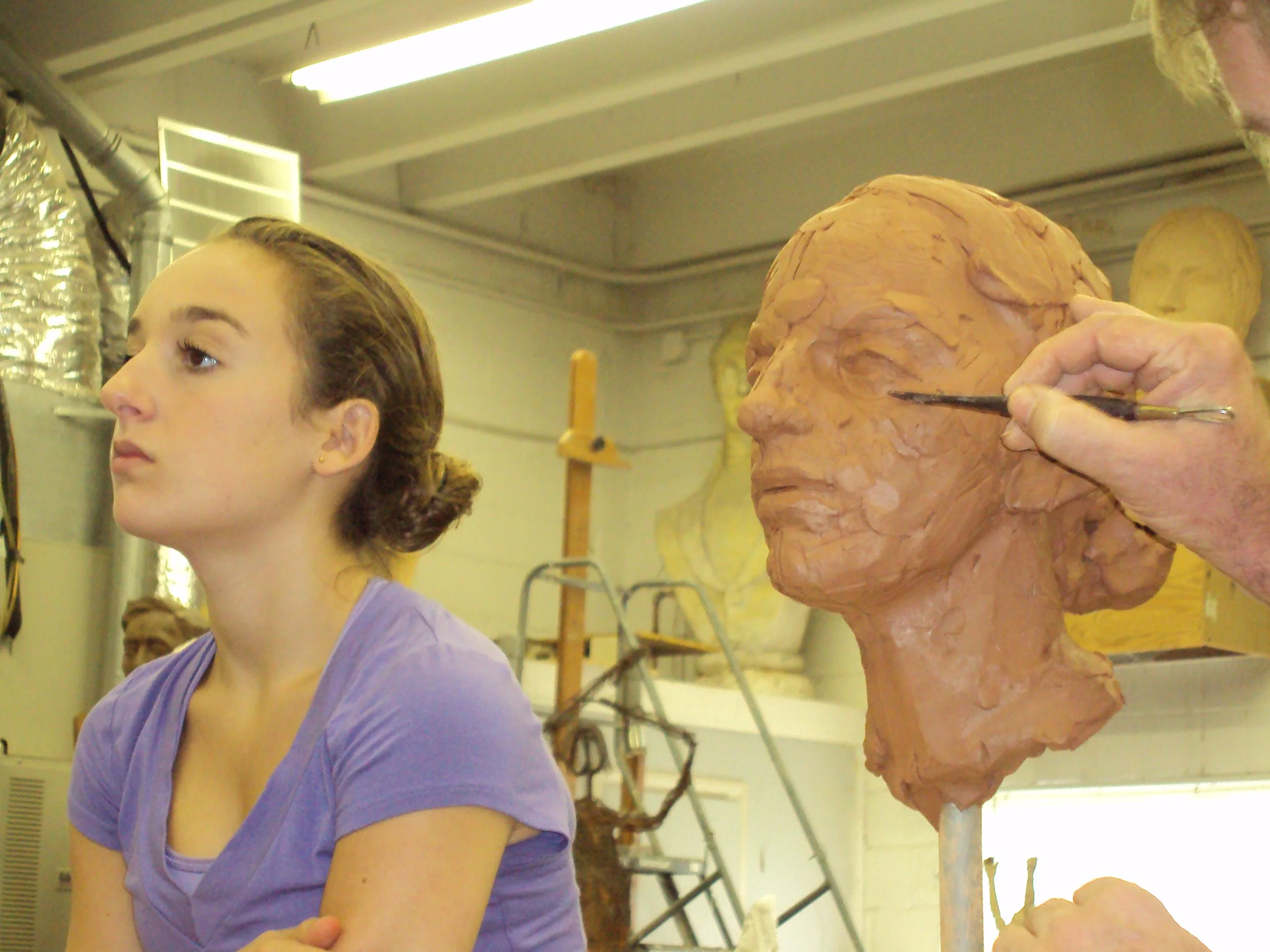 A woman with brown hair tied back in a bun is sitting in a classroom. Behind her, there is a plaster sculpture of a human head being painted by an artist. The classroom has industrial-style vents and fluorescent lighting.
