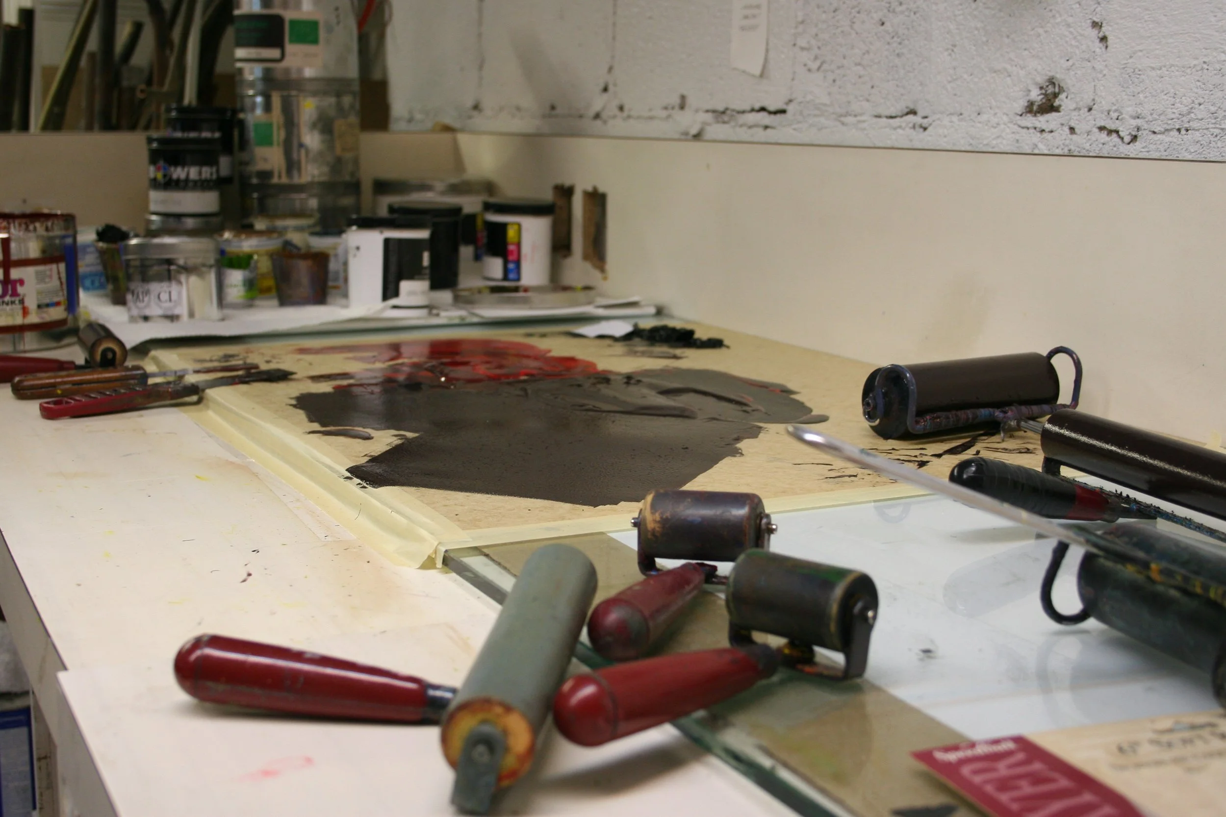 An artist's workspace with printmaking rollers, ink cans, brushes, and a etching plate covered in dark ink on a cluttered worktable.