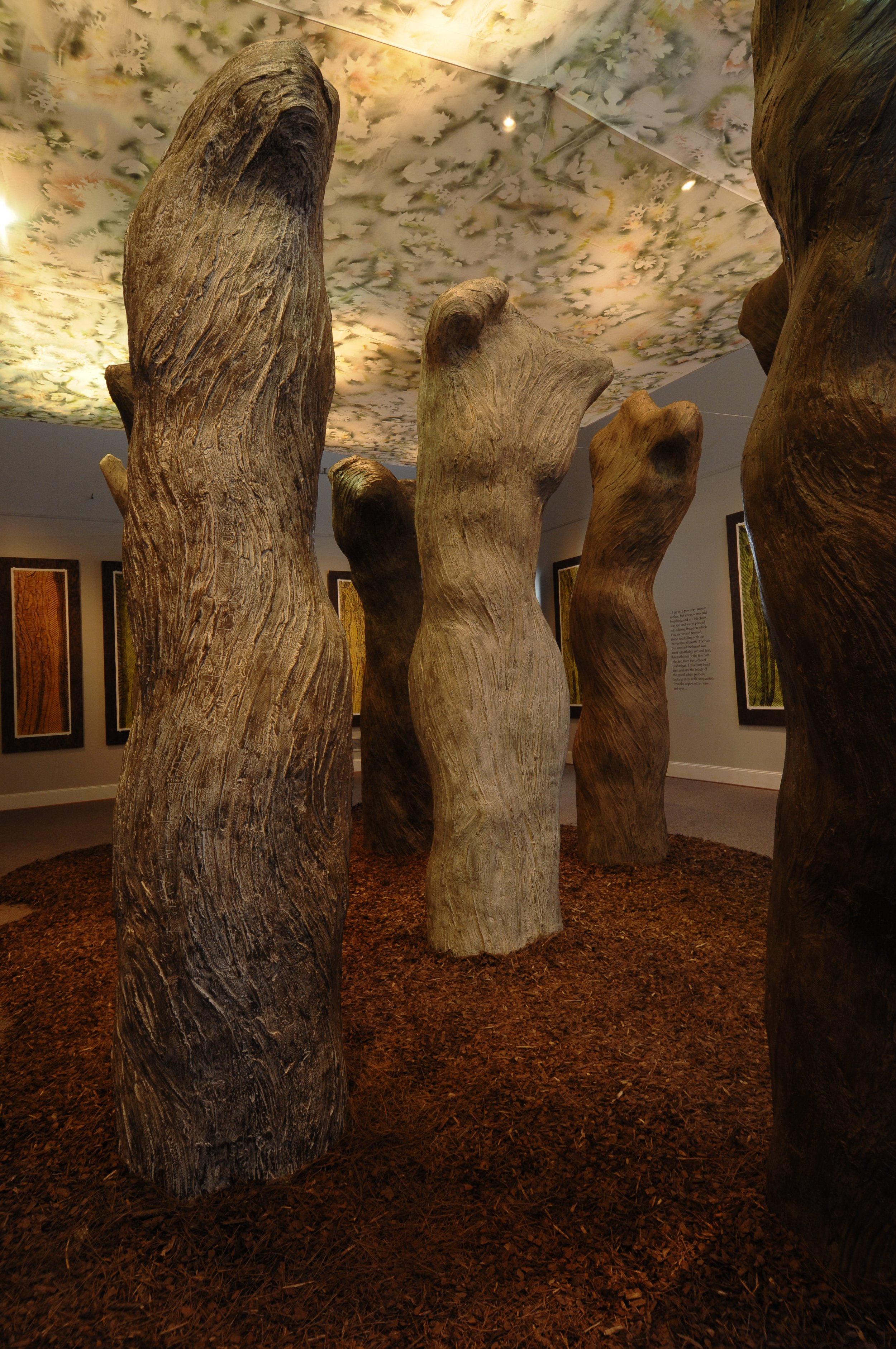 Art installation of large, upright tree trunks with textured, natural wood appearance, arranged on a ground covered with reddish-brown wood chips, in a gallery with framed prints on the walls and a ceiling with a floral pattern.
