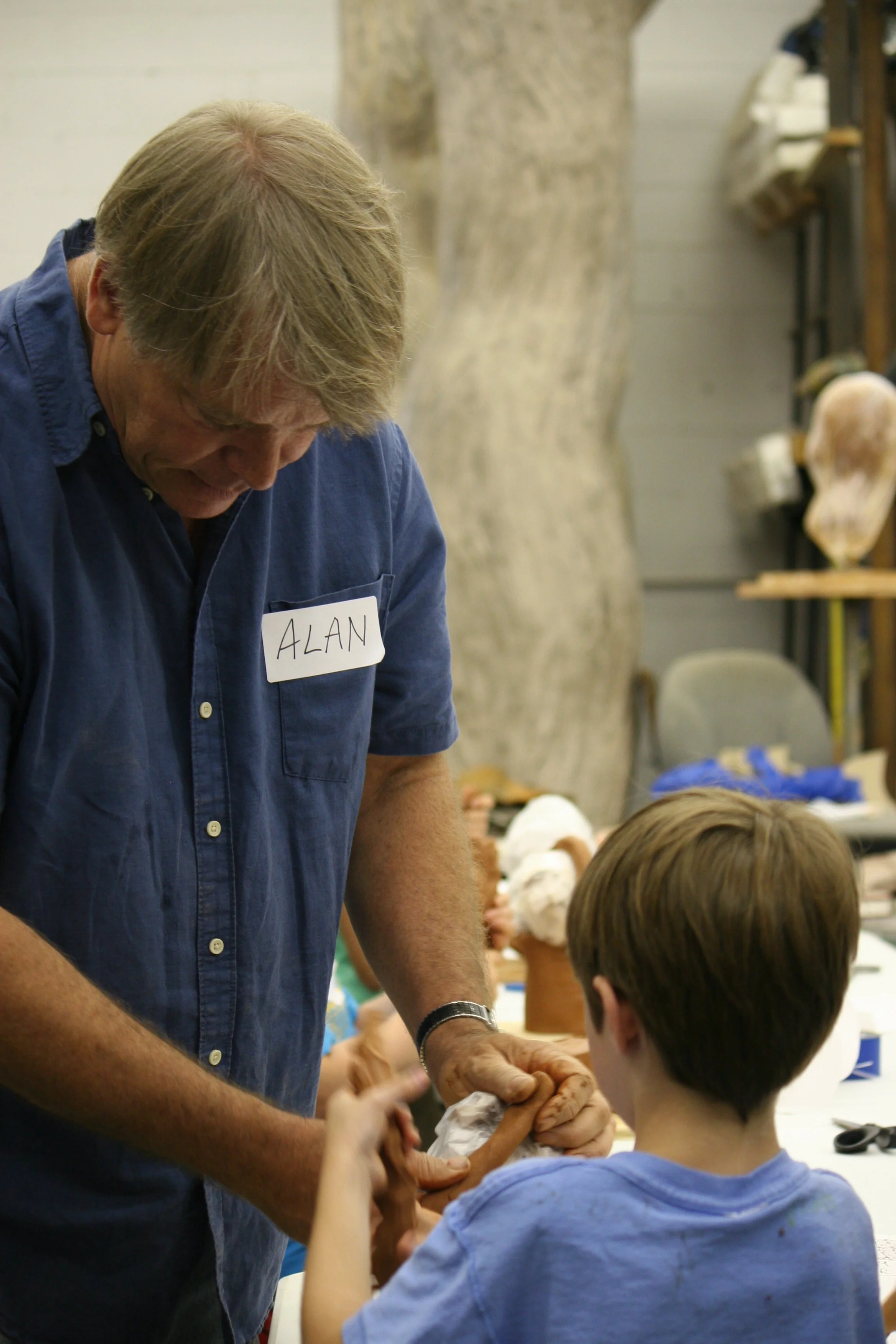 An older man with gray hair wearing a blue shirt with a name tag that says 'ALAN' interacts with a young boy in a workshop or classroom setting. The background includes shelves with various objects and other children.