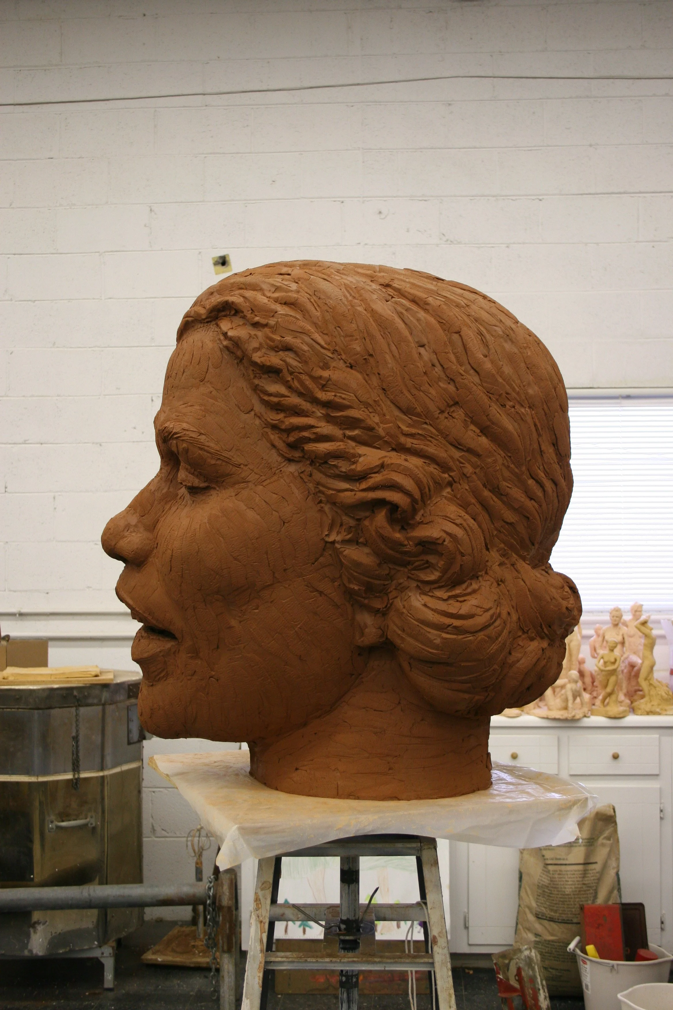 A large clay sculpture of a woman's head in profile view, positioned on a worktable in an art studio, with various art supplies and sculptures in the background.