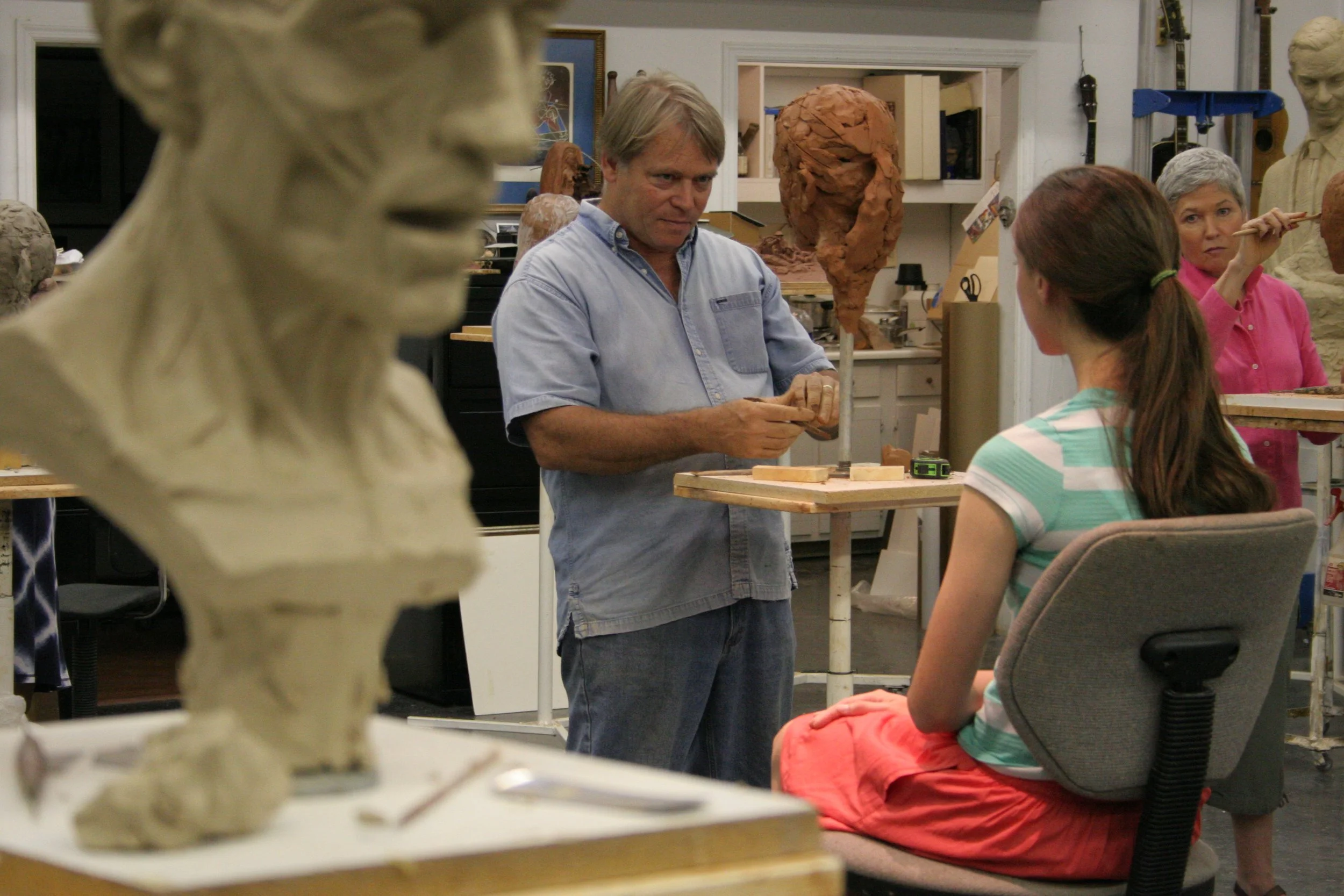 A man in a light blue shirt demonstrating pottery techniques to a woman in a striped shirt and orange pants in a pottery studio with sculptures and art supplies in the background.