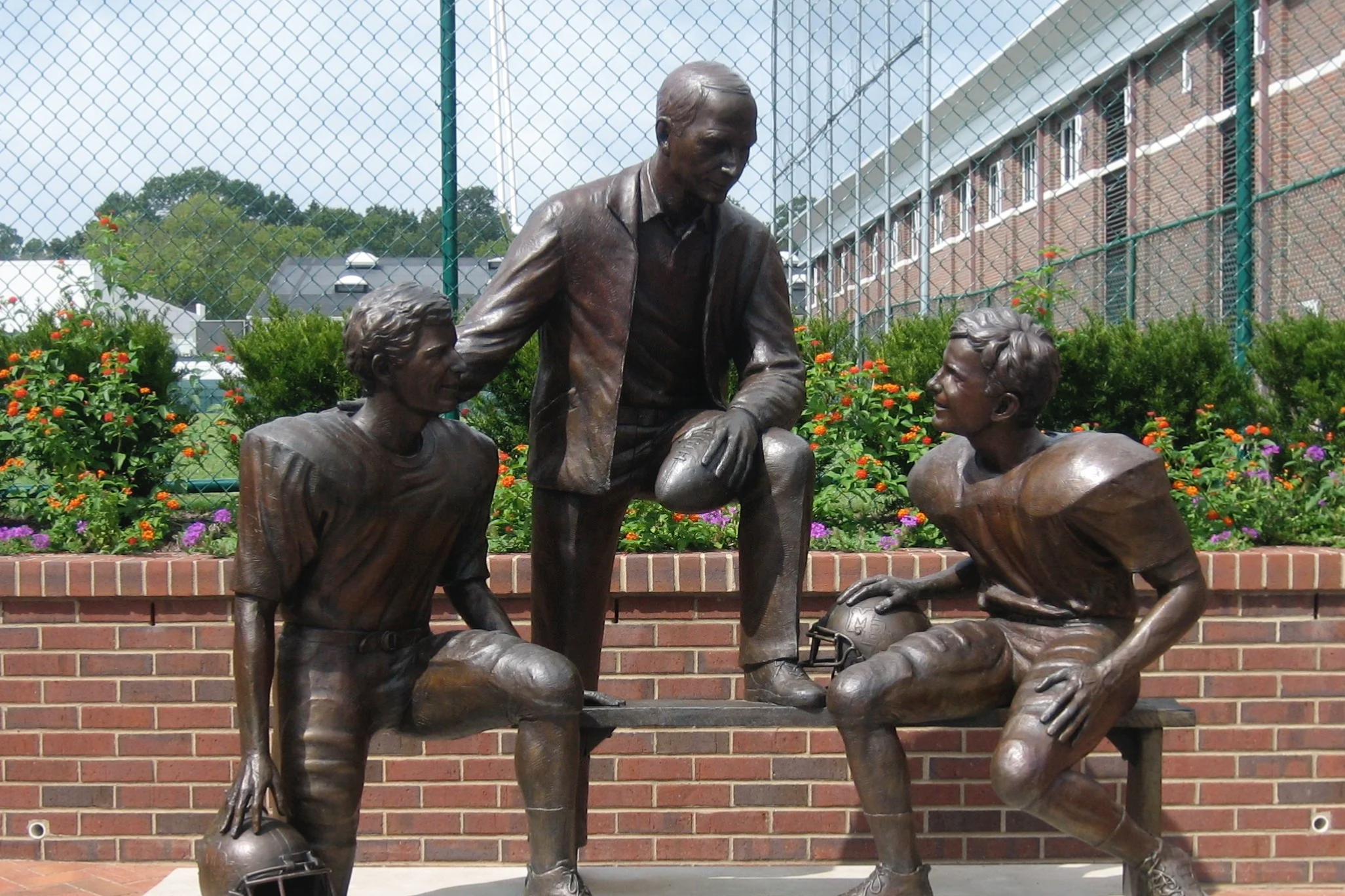 Bronze statue of three children with a football, with floral bushes and a brick building in the background.