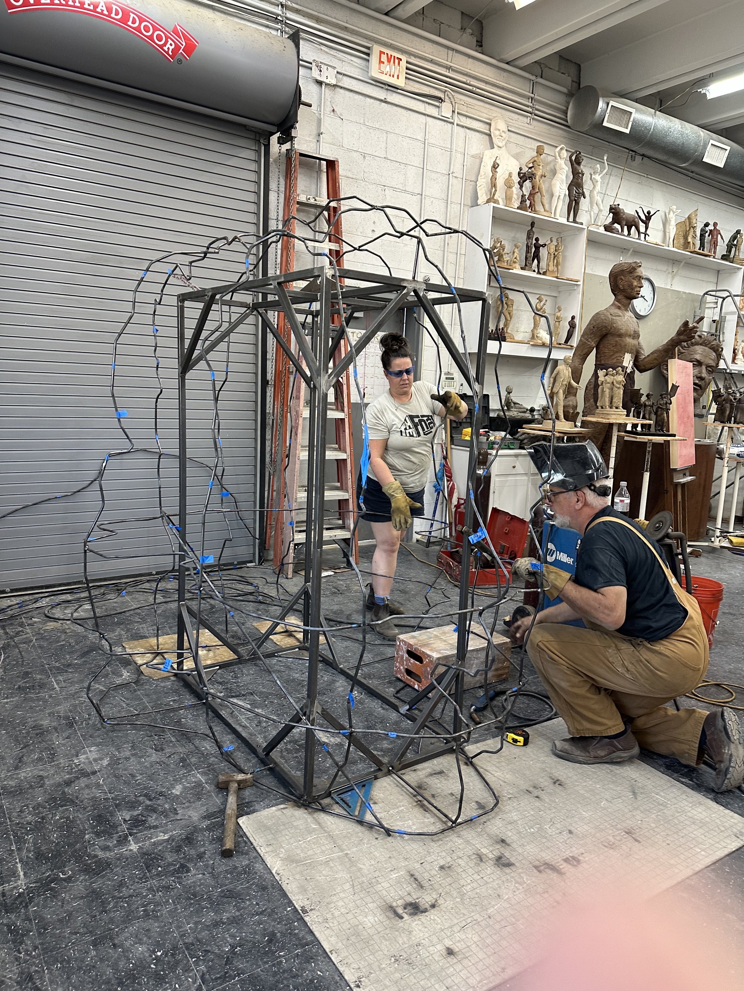 People working on a metal sculpture of a person, with a backdrop of numerous sculptures and statues on shelves in an art studio or workshop.