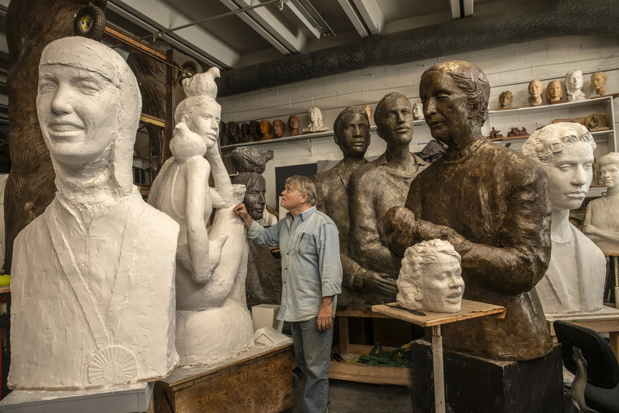 Artist working in a sculpture studio surrounded by large bust sculptures made of clay or plaster, with some finished statues in the background.