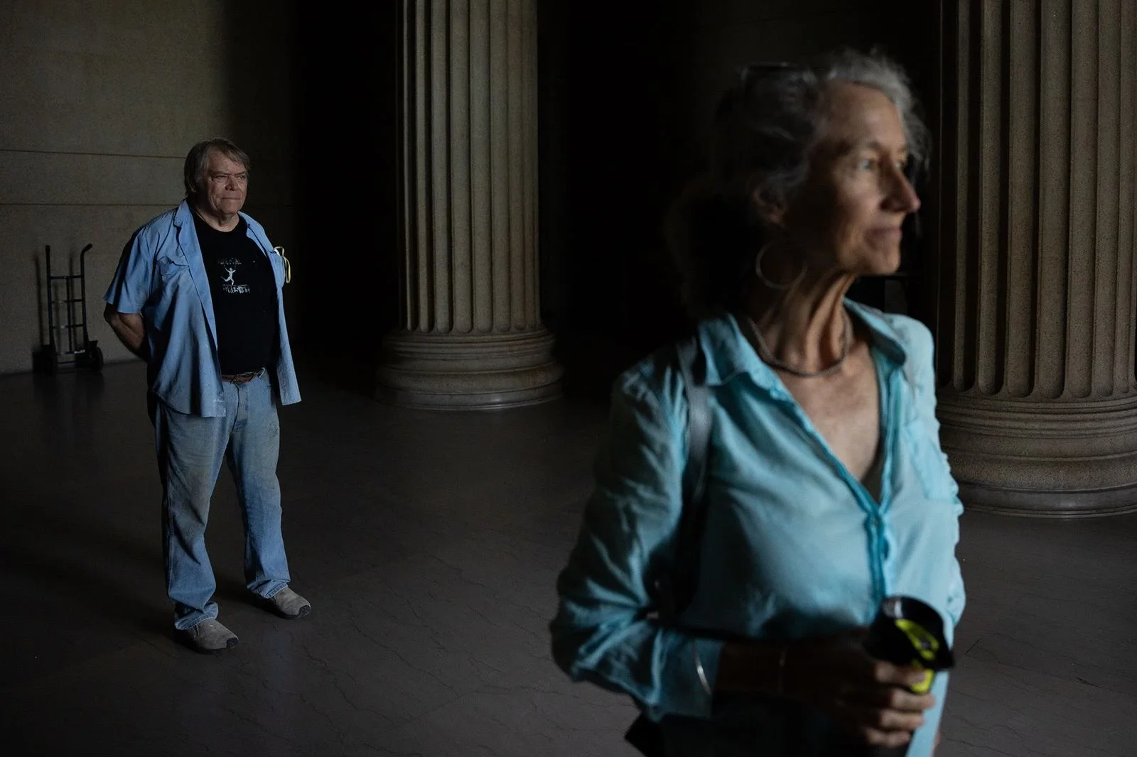 A woman in a light blue shirt holding a device facing to the right, and a man in a light blue shirt standing at a distance in a dimly lit room with large columns.
