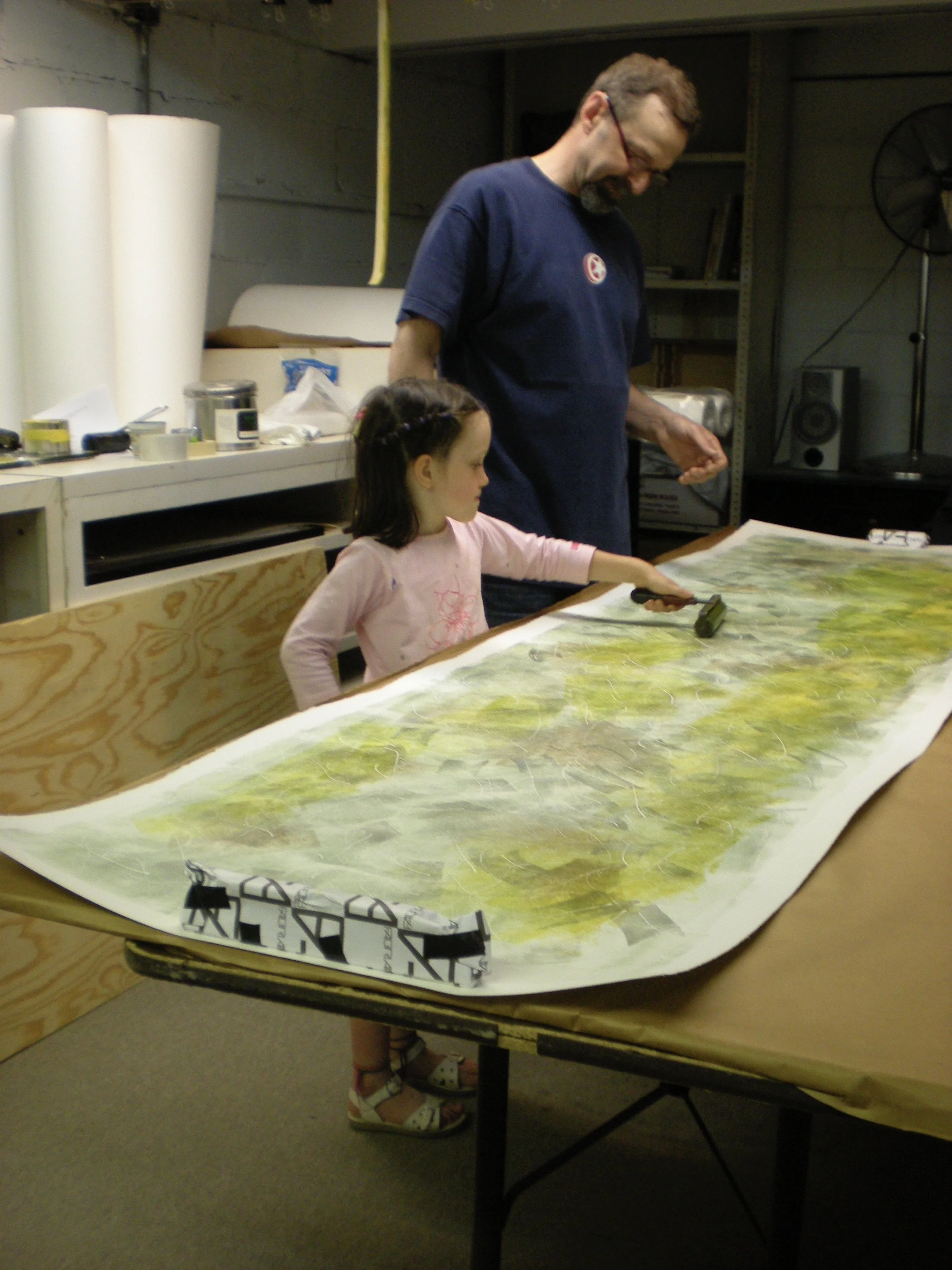 A man and a young girl working on a woodcut print on a table in a workshop or storage room. The man is standing and the girl is sitting, with various items and large white rolls in the background.