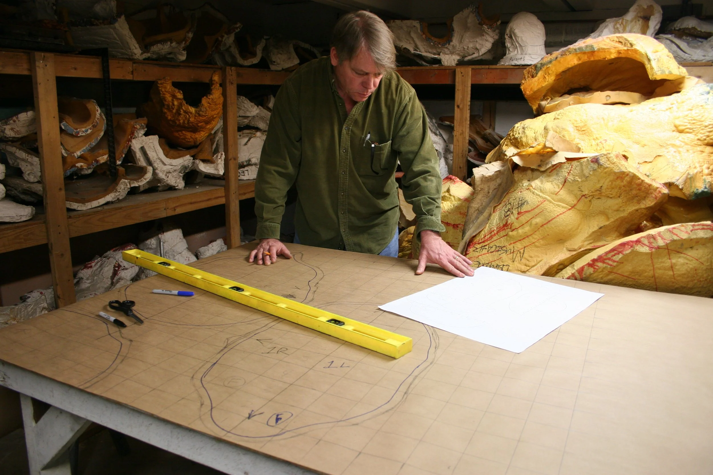 A man with gray hair wearing a green shirt is standing at a large table that has a sheet of paper with sketches and lines on it, a yellow level, pens, and scissors. In the background, there are shelves filled with large pieces of yellow, white, and b
