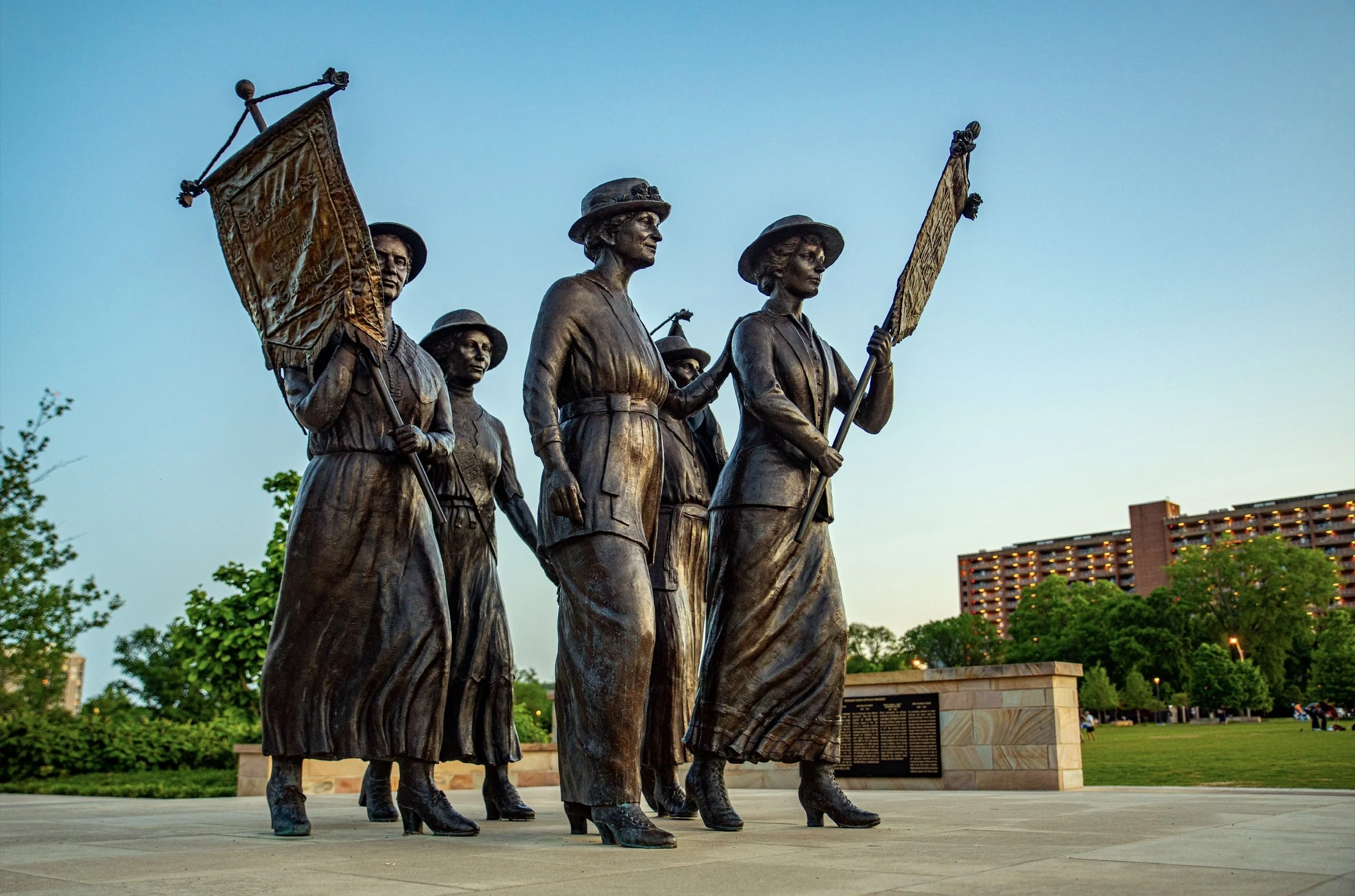 Bronze statue of women marching with flags, set in a park with trees and a building in the background at dusk.