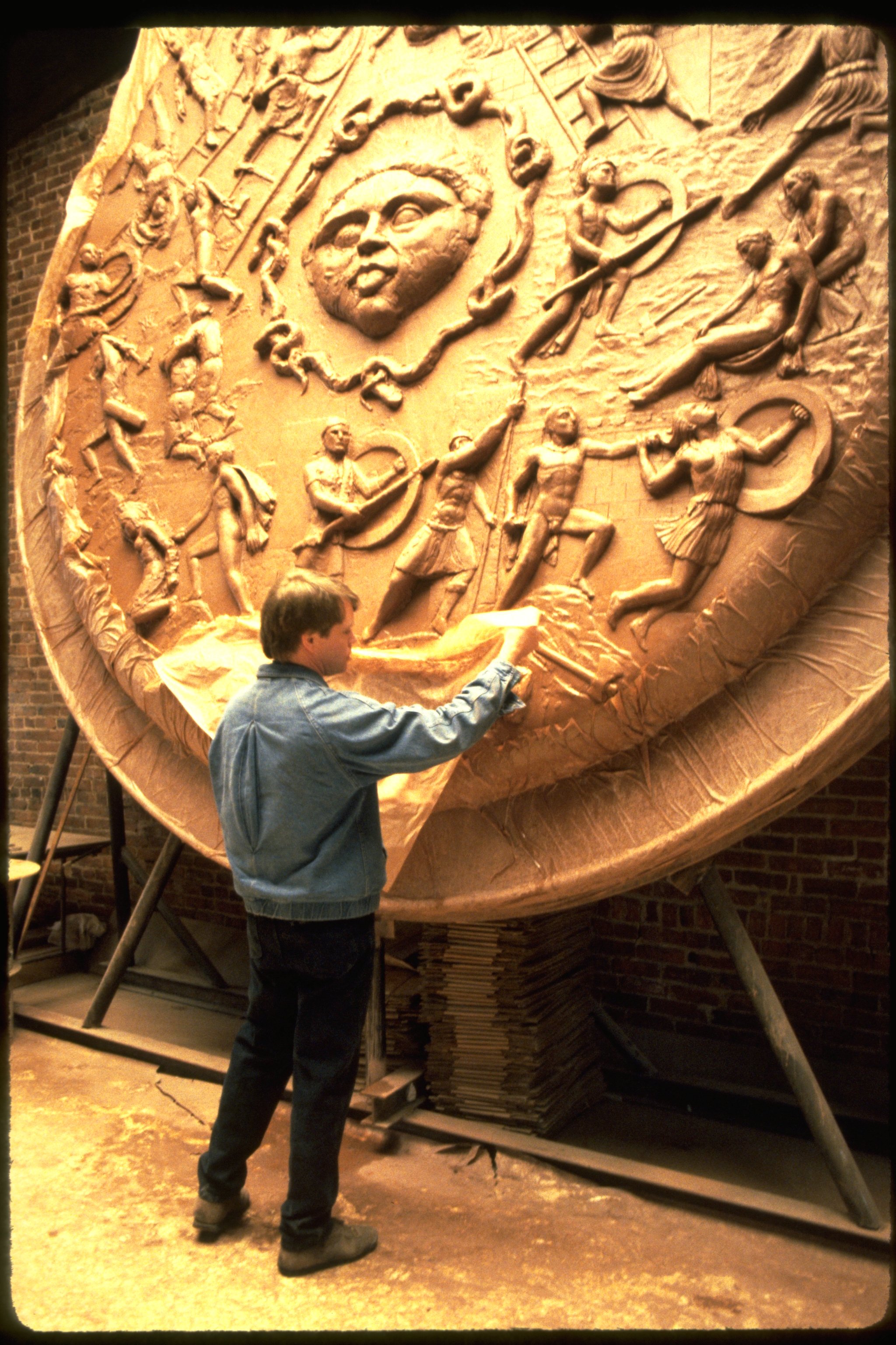 A person in a blue denim jacket and jeans is working on a large medal or relief sculpture depicting Athenas Shield with a prominent face at the top surrounded by scenes of warriors and people, mounted on a stand in a workshop.