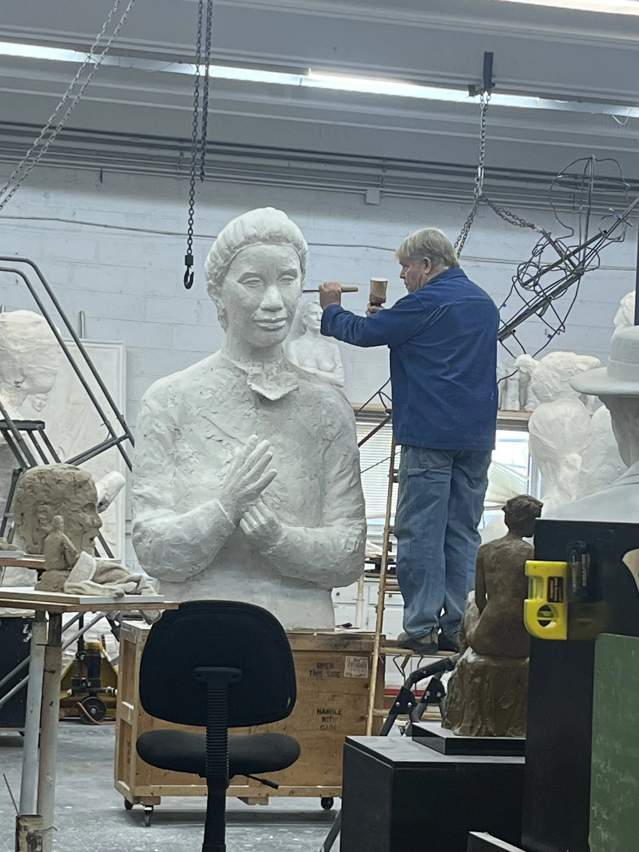 A person working on a large white sculpture of a woman's head in an art studio, with various smaller sculptures and art supplies around.