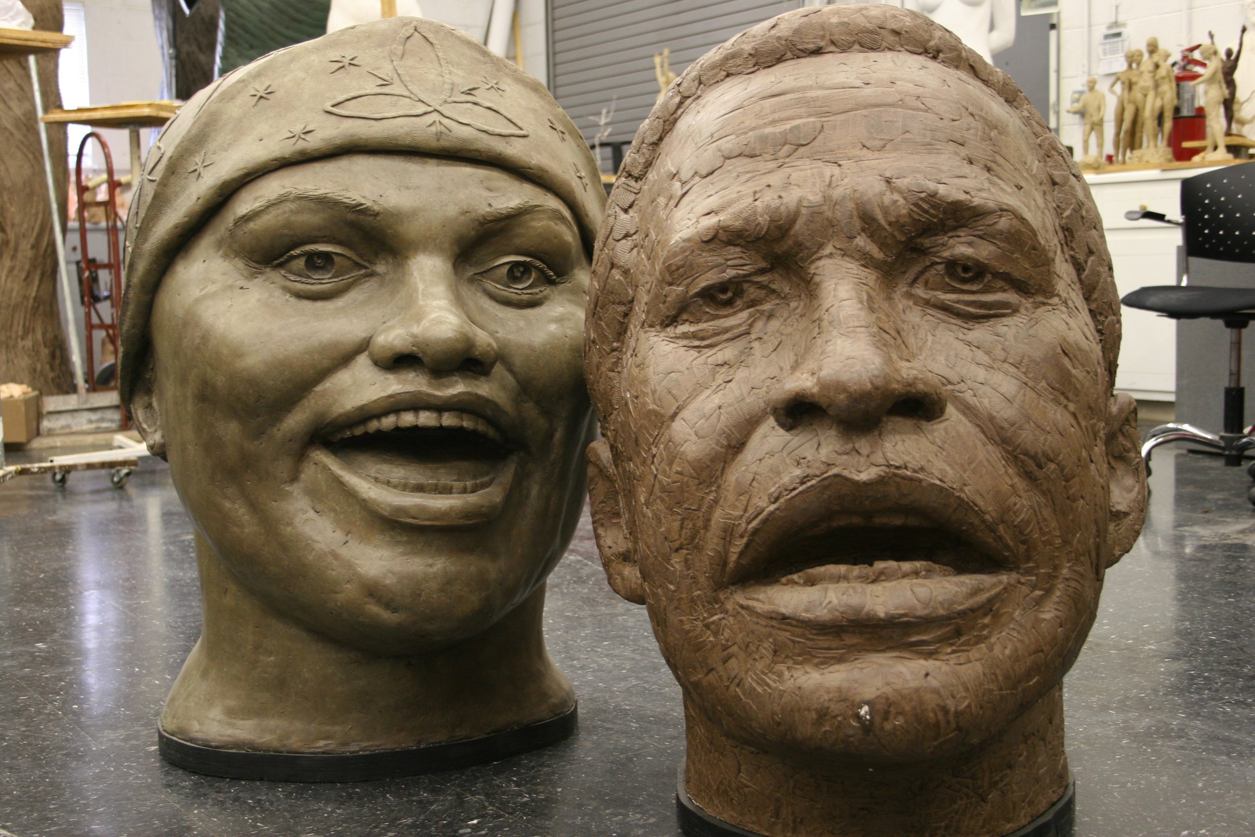 Two large artisan sculptures made of  or similar material, one depicting a smiling woman and the other a serious face with textured features, set indoors in a studio with various art tools and sculptures in the background.  Cultural Heroes
