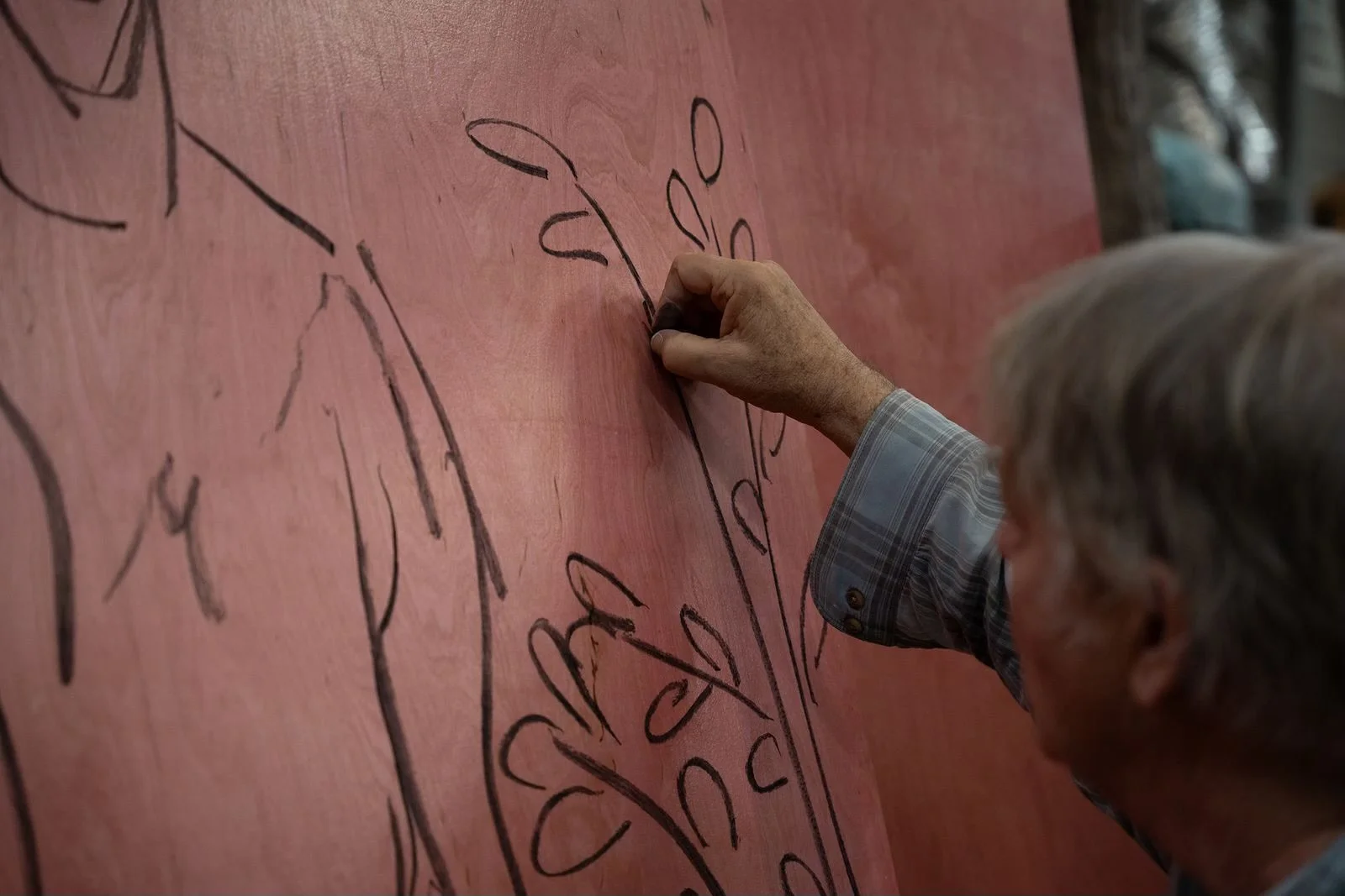 Close-up of an elderly man's hand drawing on a large pink canvas with a black marker, with sketchy floral outlines visible.