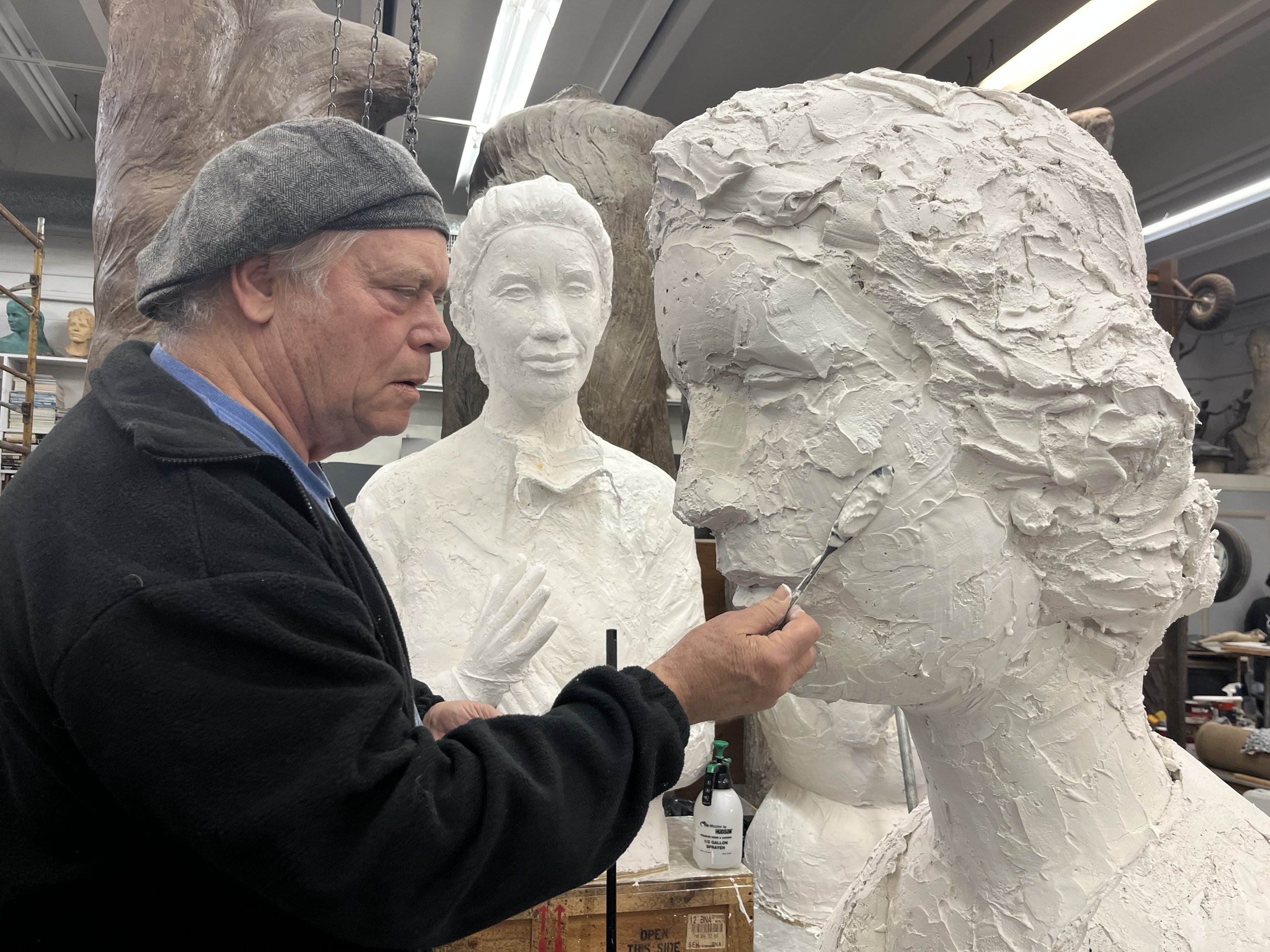 An artist wearing a grey beret and black jacket sculpting a white plaster bust of a woman in a workshop filled with sculptures and art supplies.