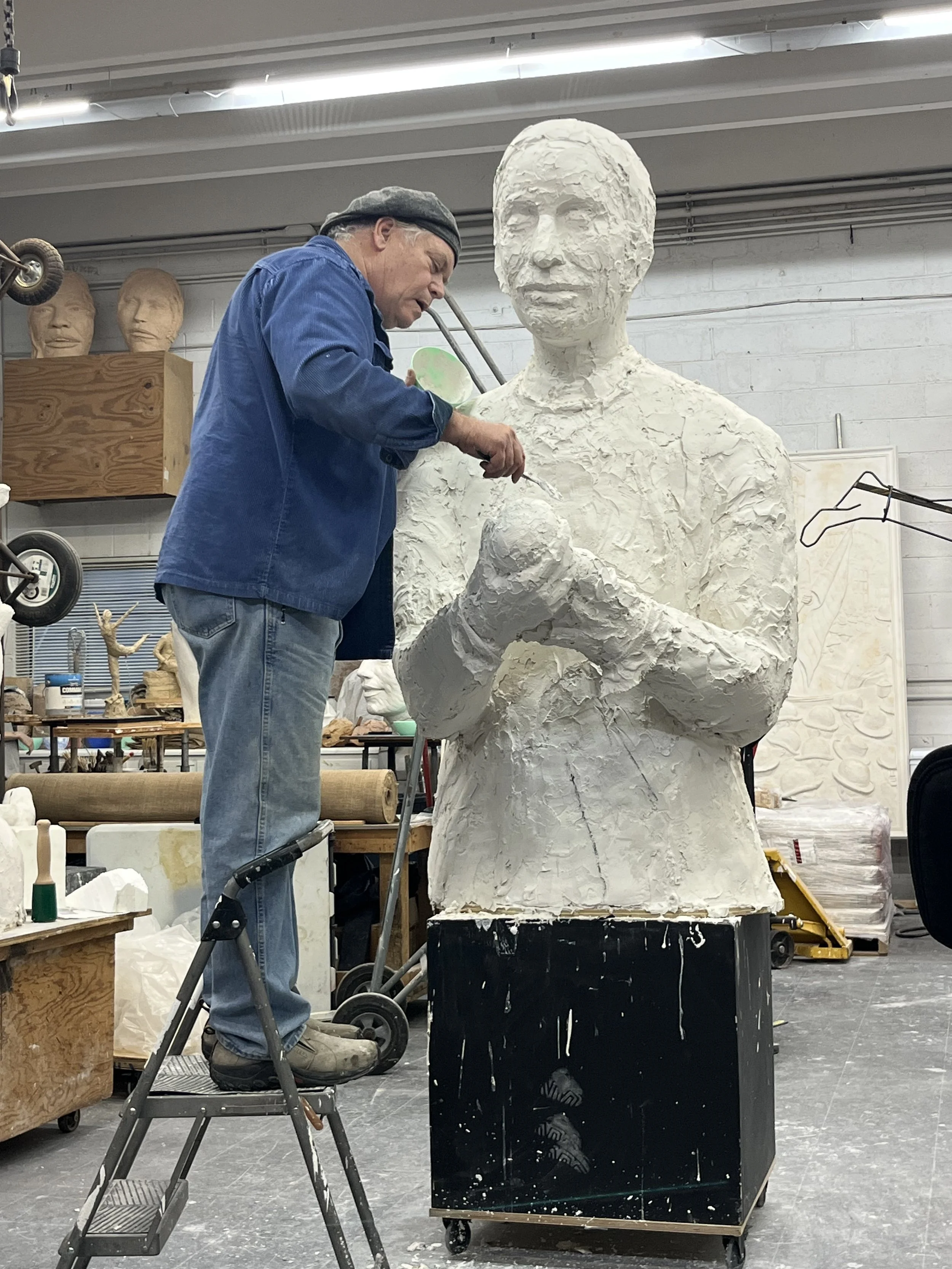 An artist working on a large, partially completed sculpture of a person’s bust in a workshop.