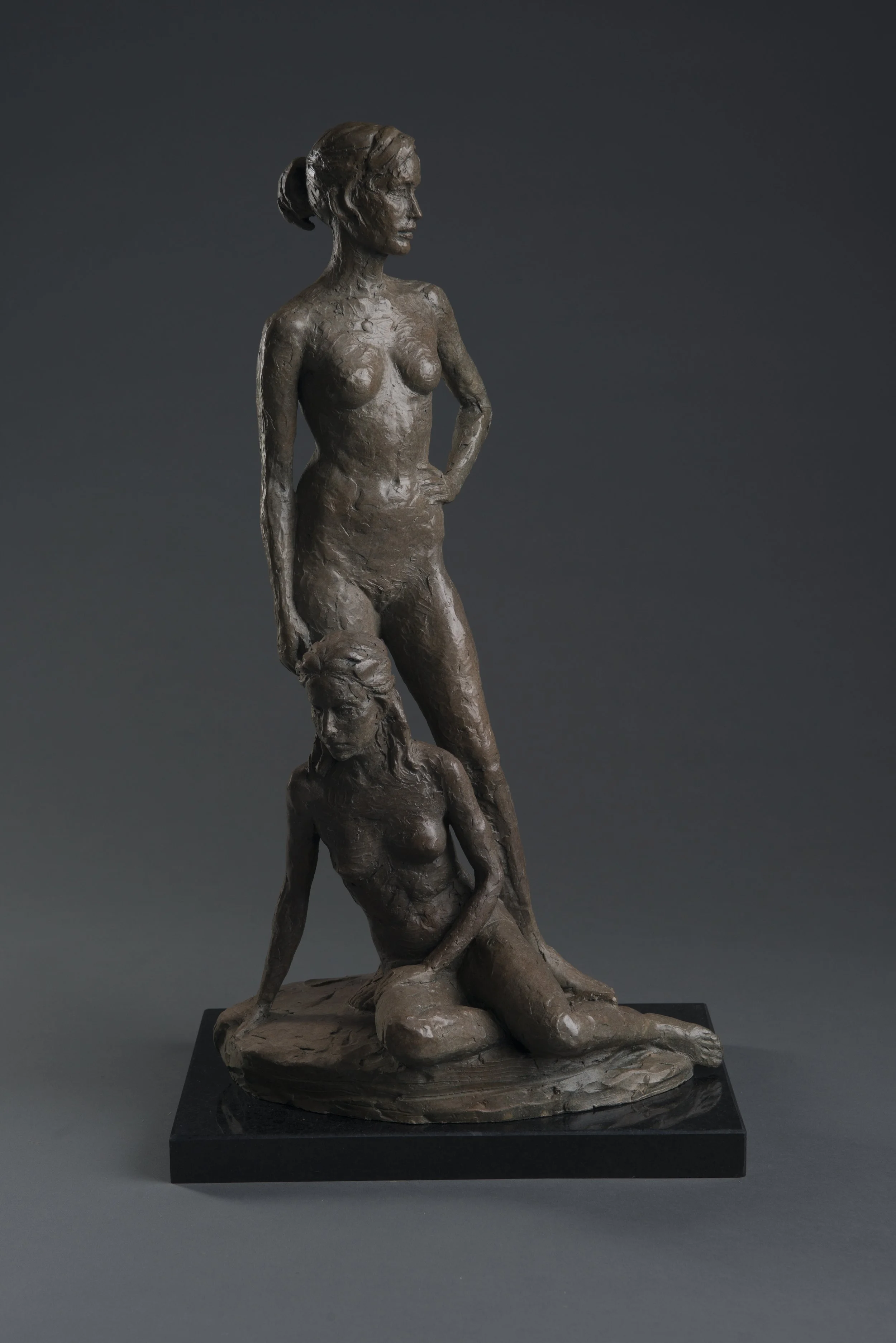 Bronze sculpture of a woman standing next to another woman, with one hand on her hip, set against a dark gray background.