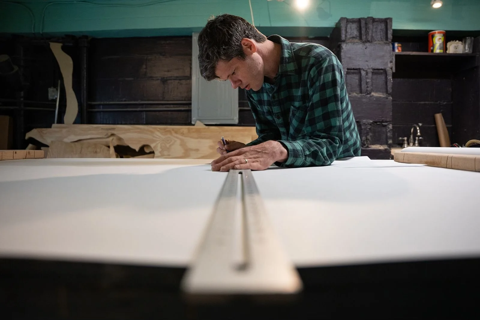 A man in a green plaid shirt measuring and marking wood on a large table in a woodworking shop.