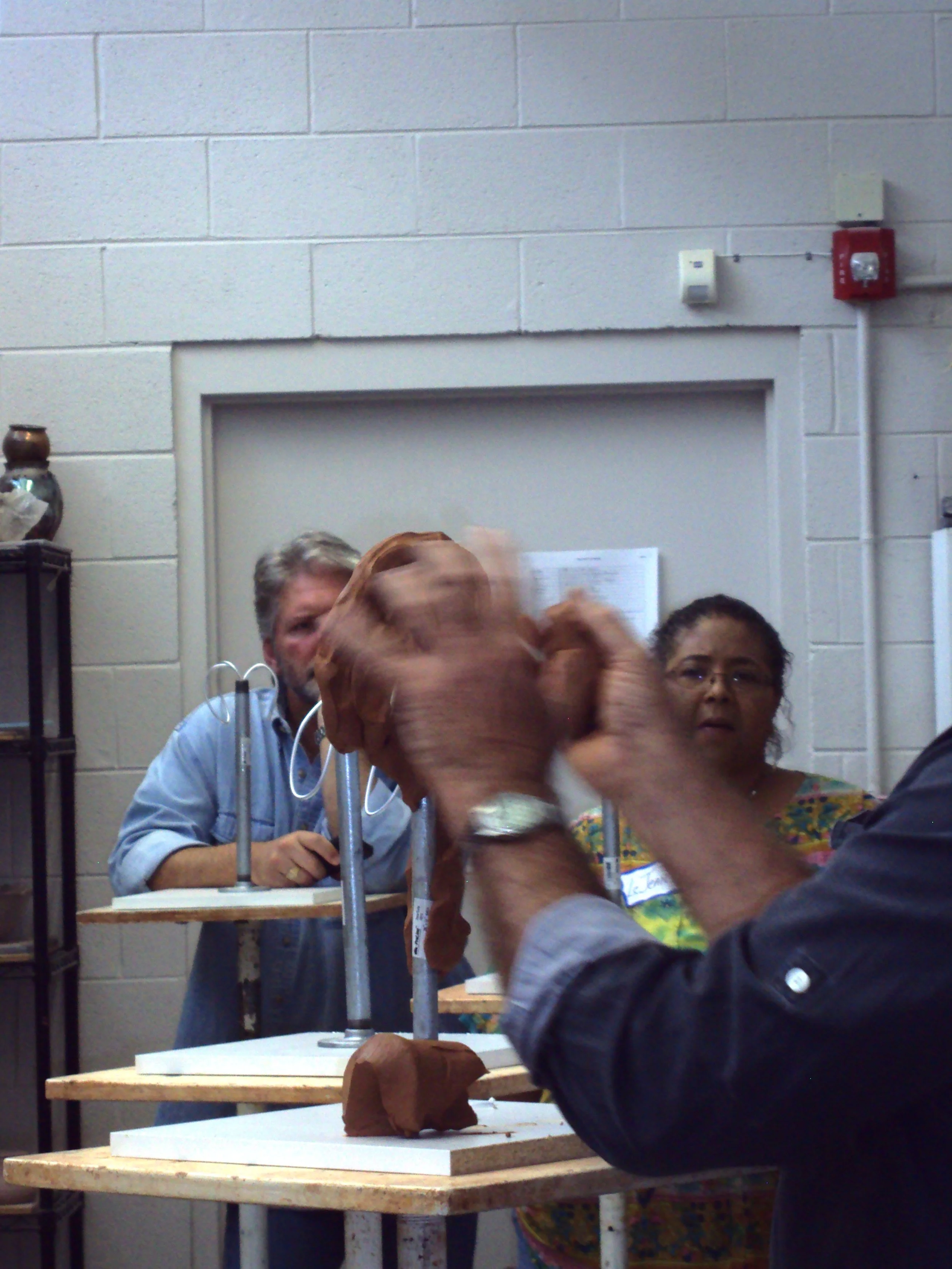 People participating in a clay modeling activity in a classroom with beige cinder block walls. A woman and a man are visible in the background. Several individuals' hands are working on a brown clay sculpture placed on paired white and wooden tables.