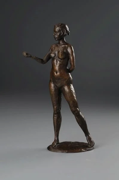 Bronze sculpture of a standing female figure with short hair, holding an object in her right hand, against a plain gray background.