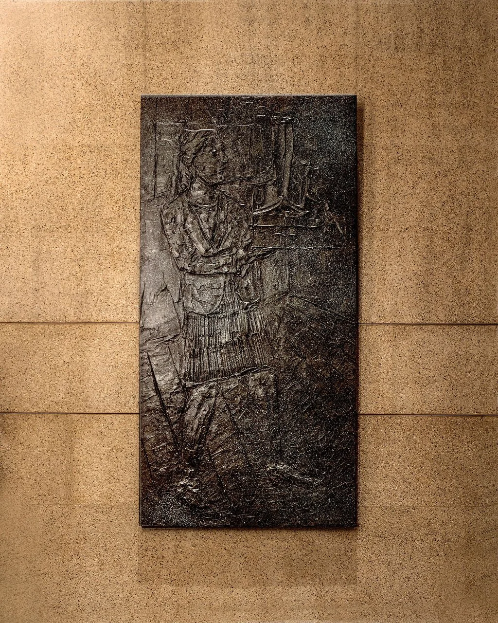 A bronze relief sculpture of a woman architect standing with her arms holding a building model,  on a beige wall.