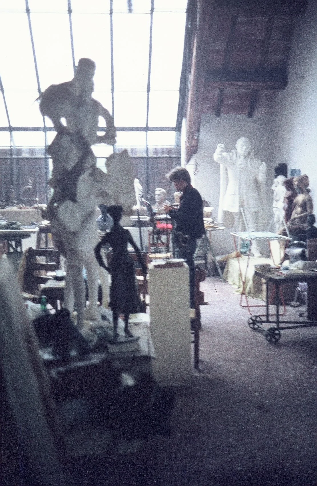 Art studio filled with sculptures, art supplies, and a person working on a piece, with large windows and sculptures in the background.