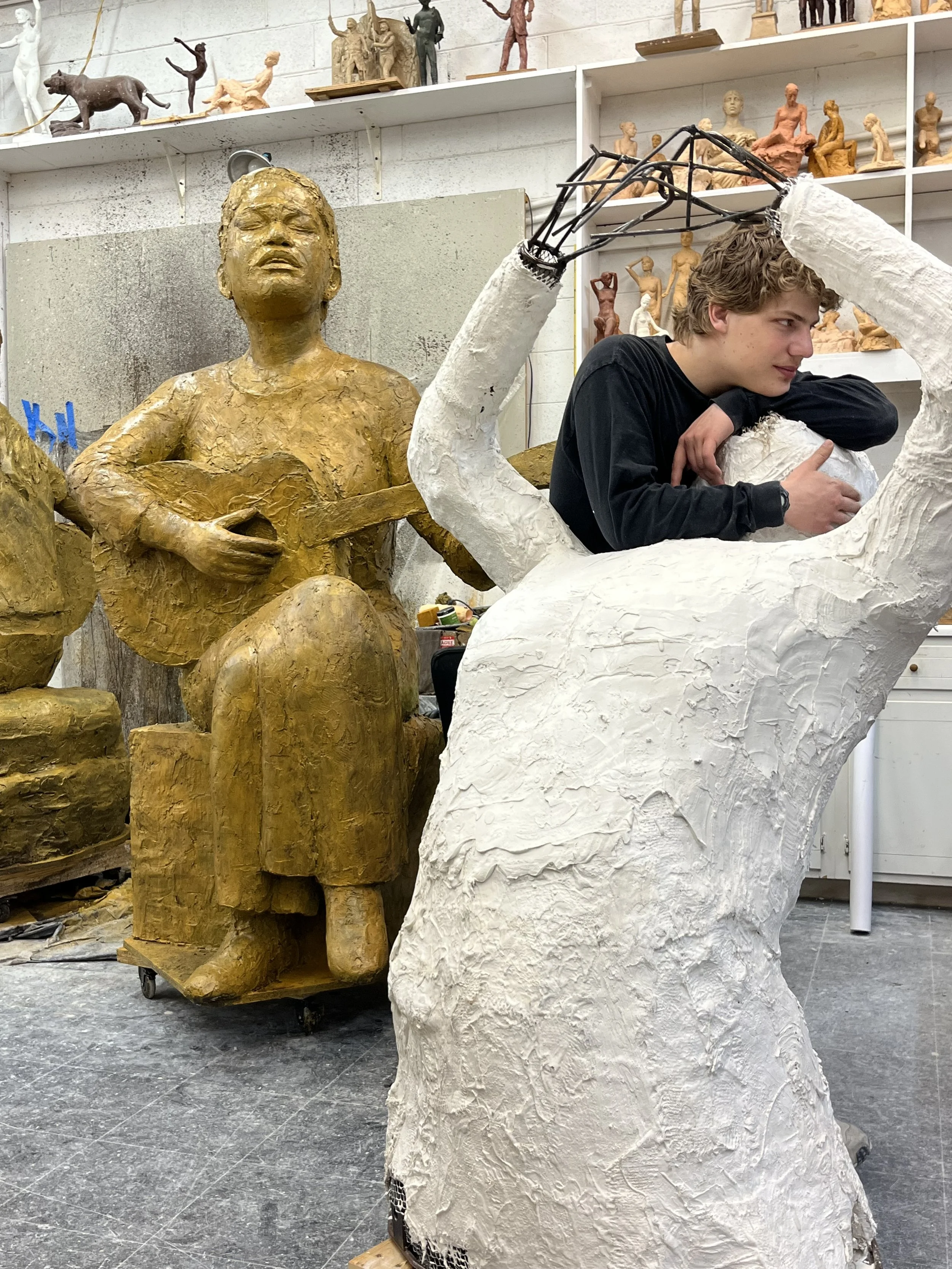 A young man working on a sculptural art piece in a studio, with a partially finished white sculpture of a seated figure and a gold-colored sculpture of a woman playing a guitar nearby.