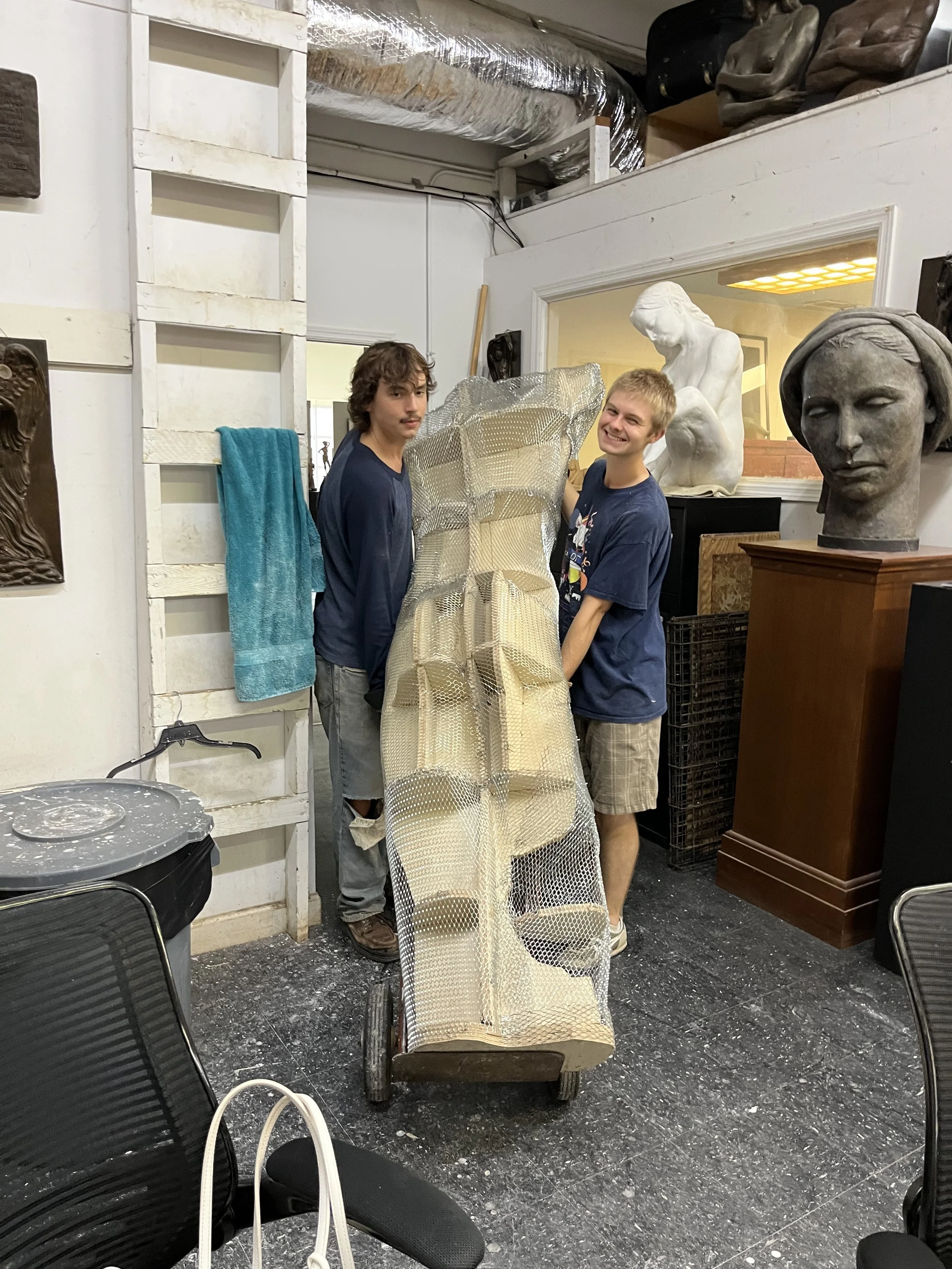 Two young men standing next to a sculpture in an art studio. One is looking at the camera with a slight smile, the other is looking at the sculpture. The studio has various sculptures and art supplies.