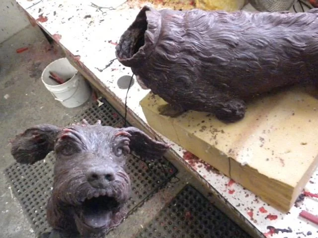 A dog in a workshop with its head tilted up, looking at the camera with tongue out, next to a large wooden sculpture of a dog on a worktable.