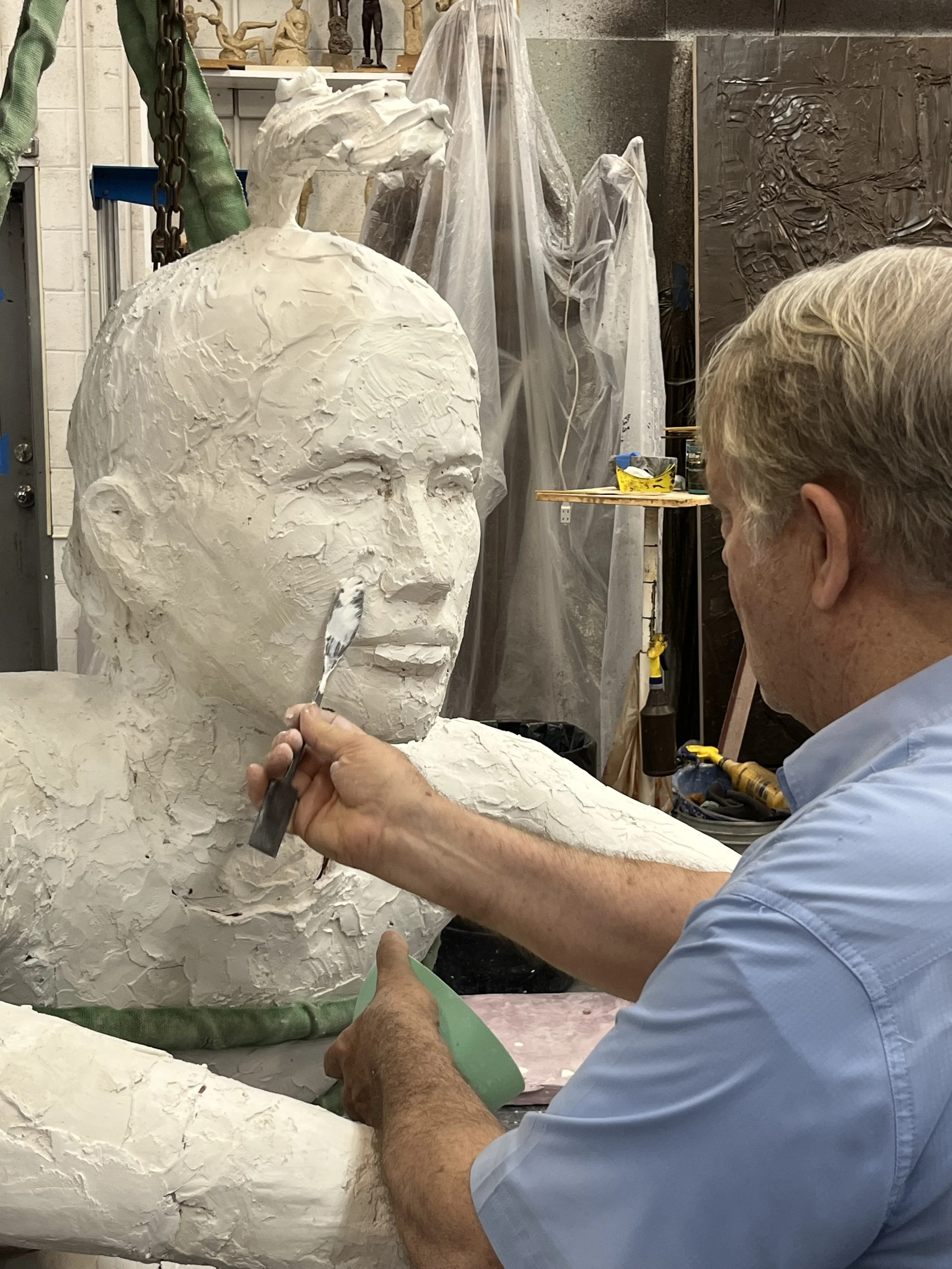 An artist creating a large sculpture of a person's face with white material, working with sculpting tools in a studio.