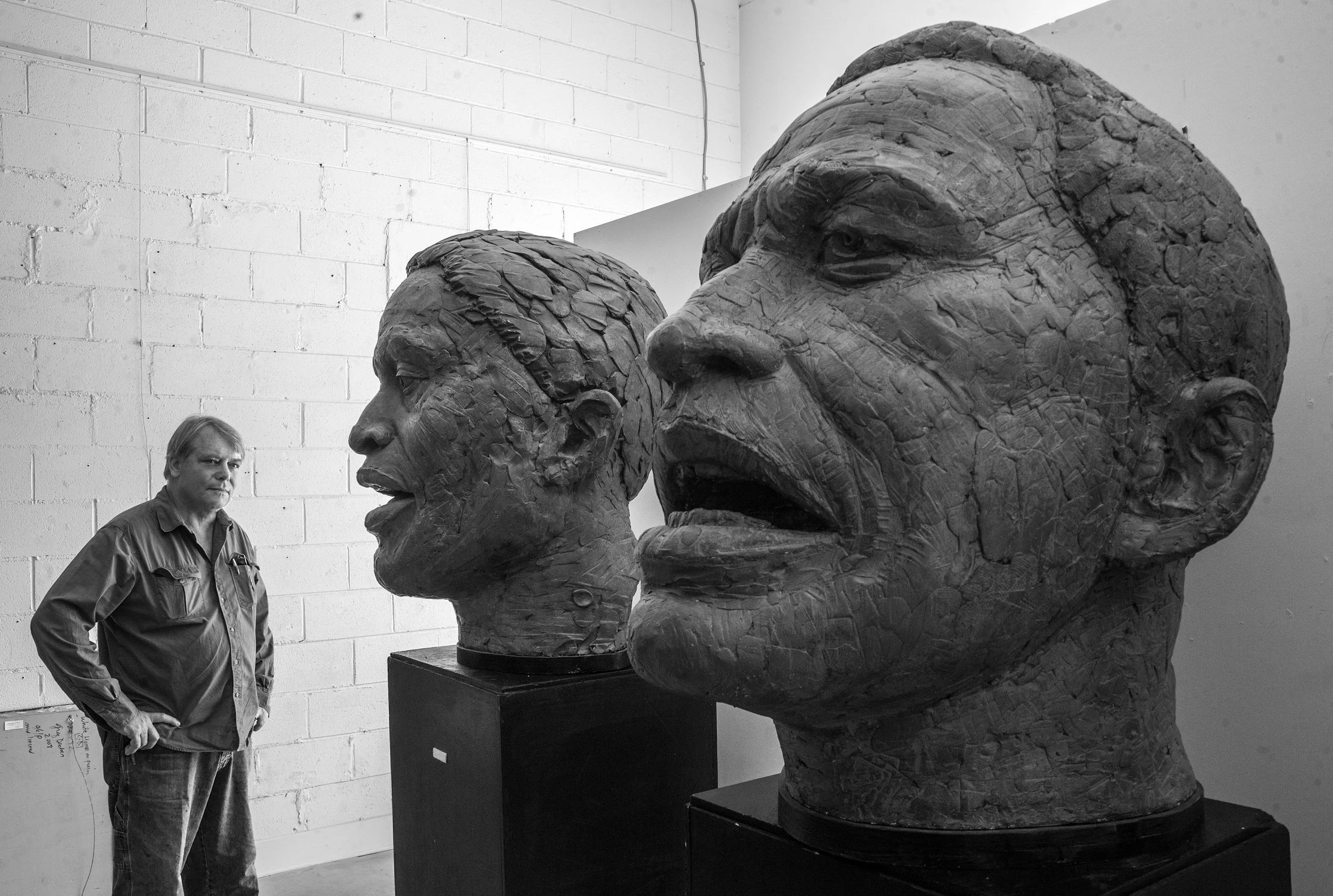A man stands with his left hand on his hip, looking at large sculptures of human faces in a room with white brick walls.