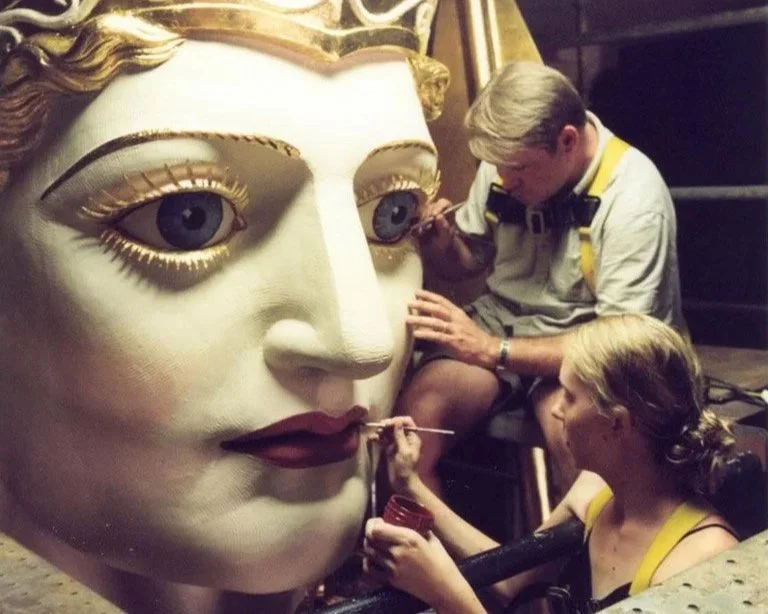 Two artists painting a large, realistic face sculpture of a woman with blonde hair and blue eyes.