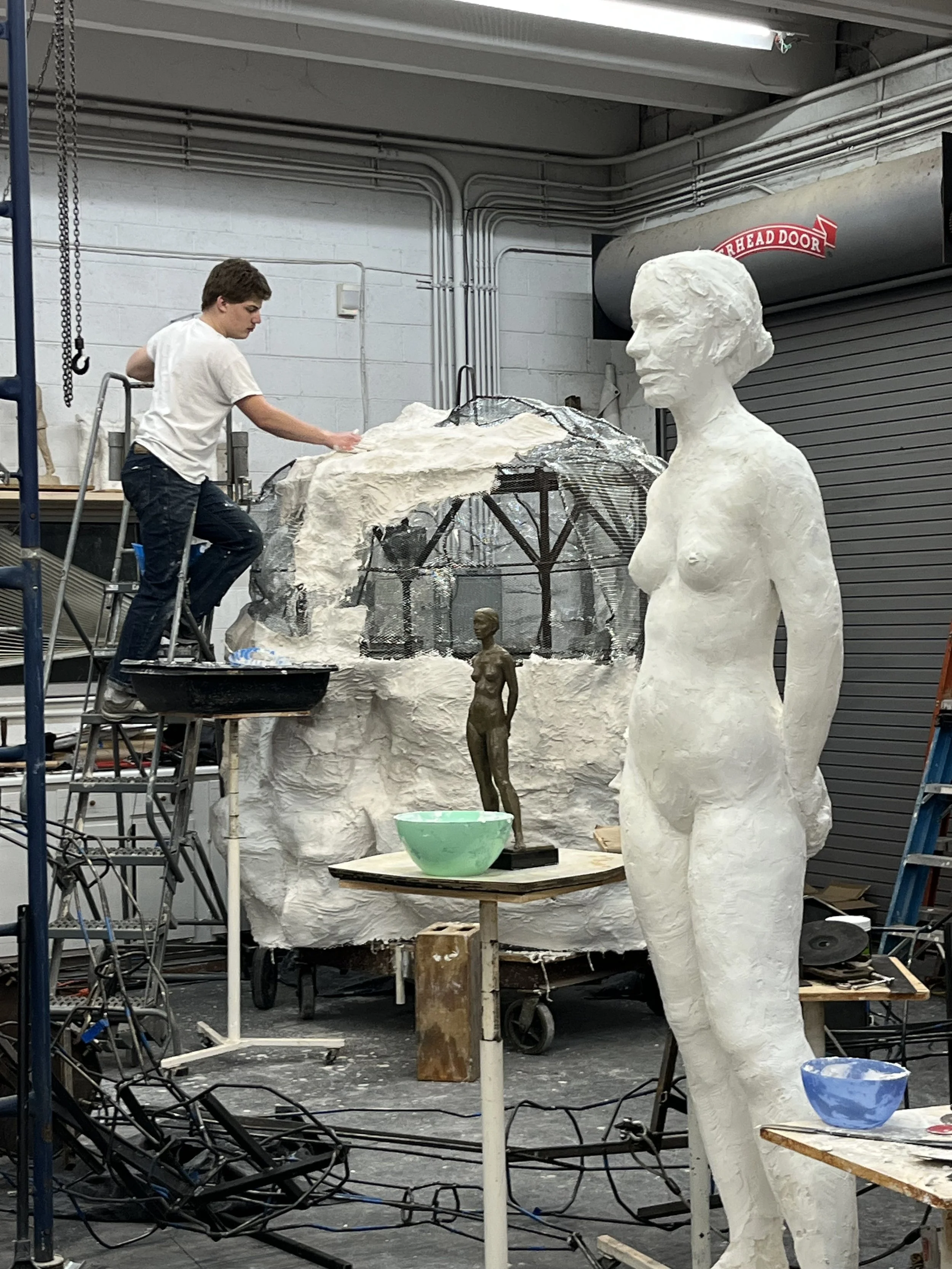An artist working on a sculpture studio with a large white sculpture of a woman, a small bronze figure, and various art supplies and equipment.
