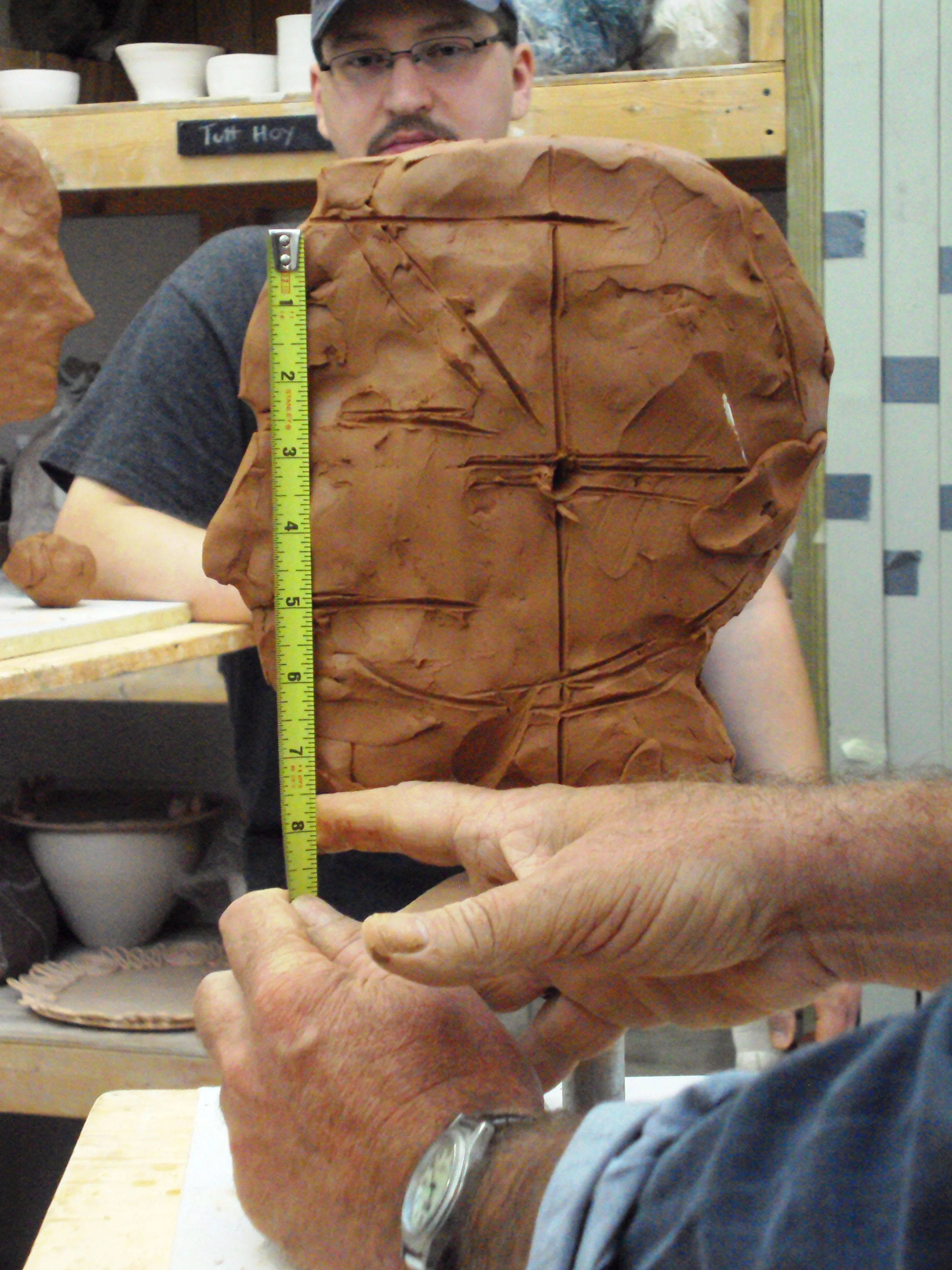 A person holding a large piece of clay or similar material, with a measuring tape showing approximately 5 inches in height, in a workshop or studio setting.