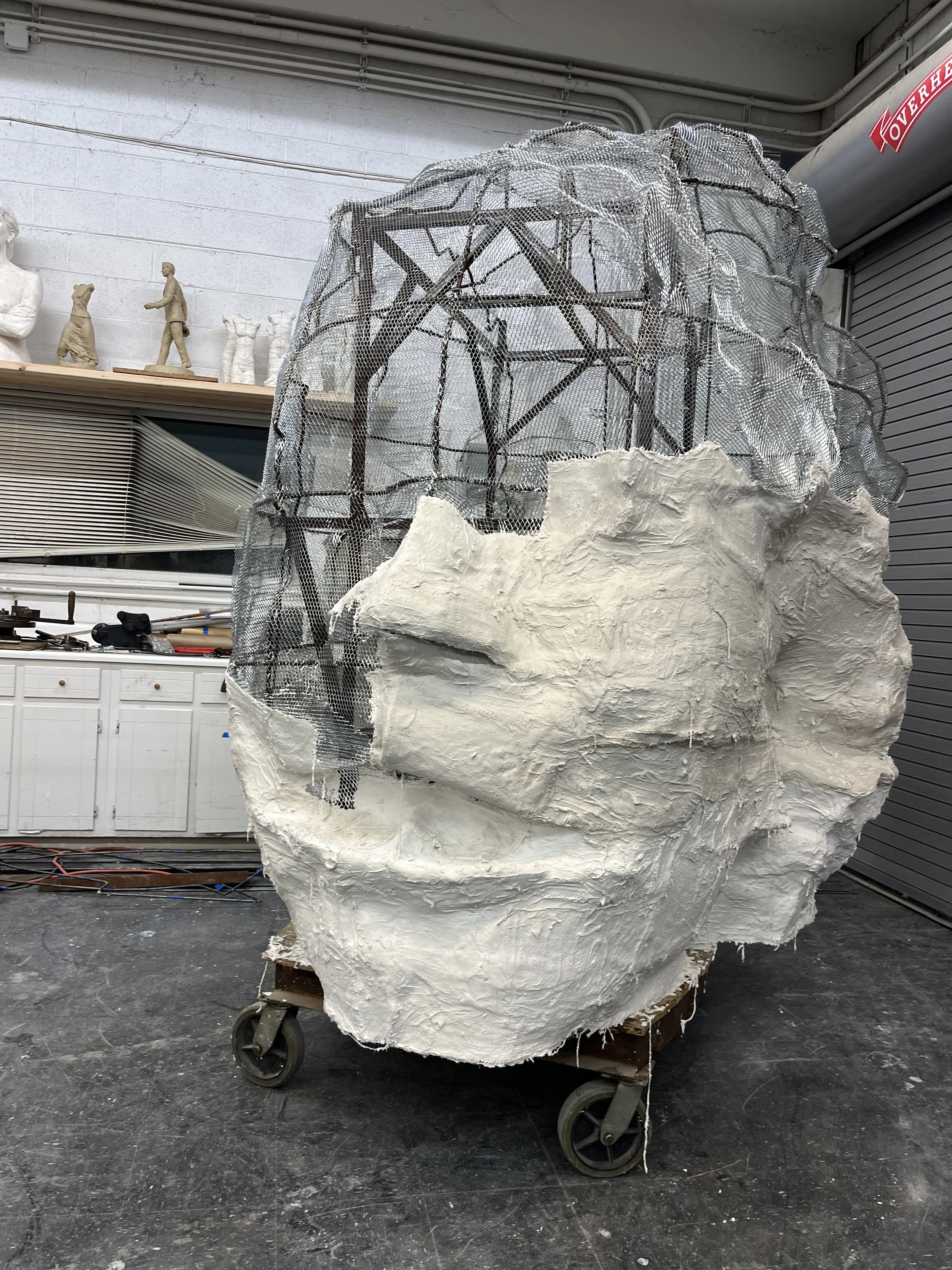 Studio art sculpture of a large white rock formation on a wheeled platform, with metal framework and protective mesh.