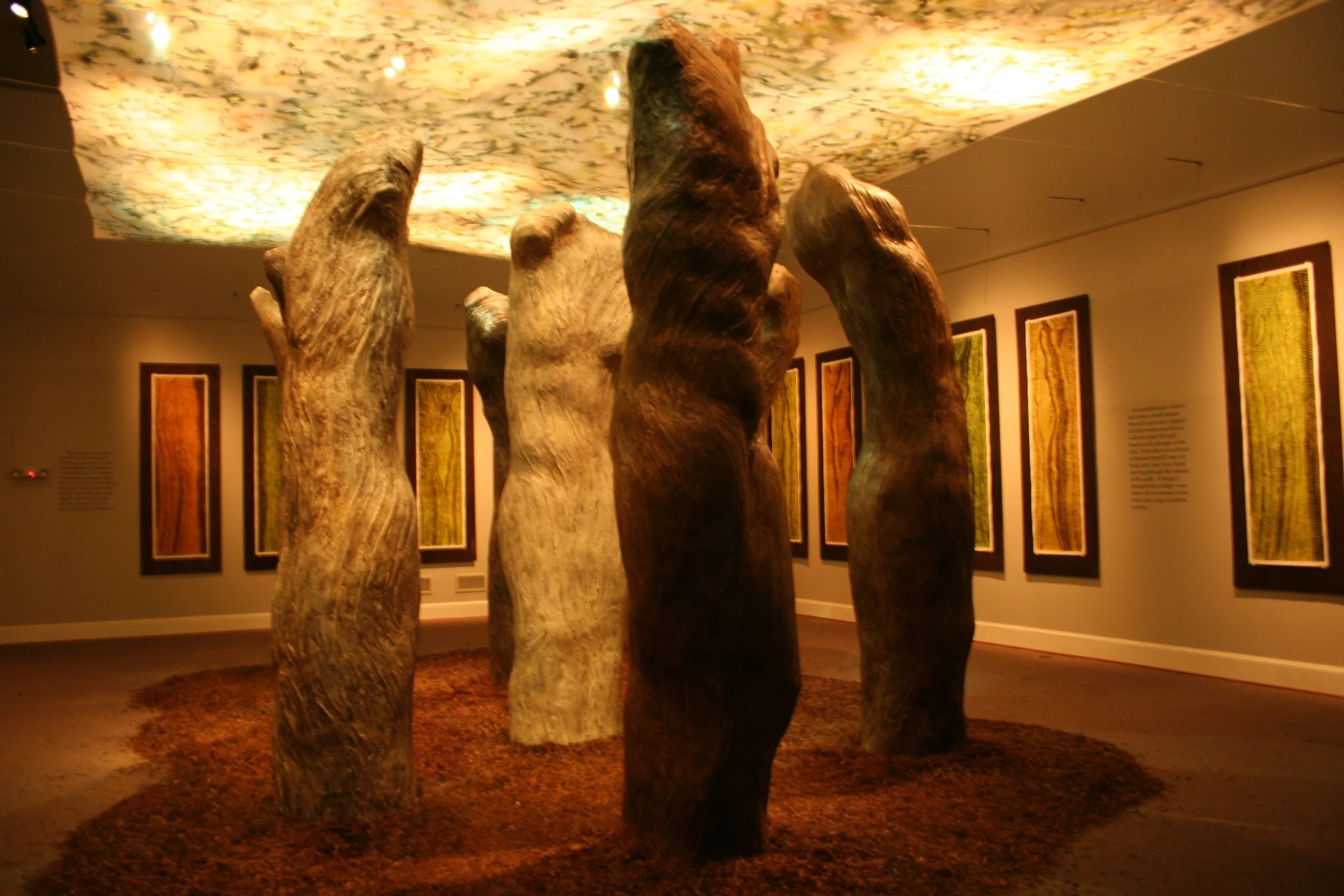 An art exhibit in a gallery featuring tall, abstract wooden sculptures resembling human forms, with framed paintings or prints on the walls behind them. Dream Forest Installation.